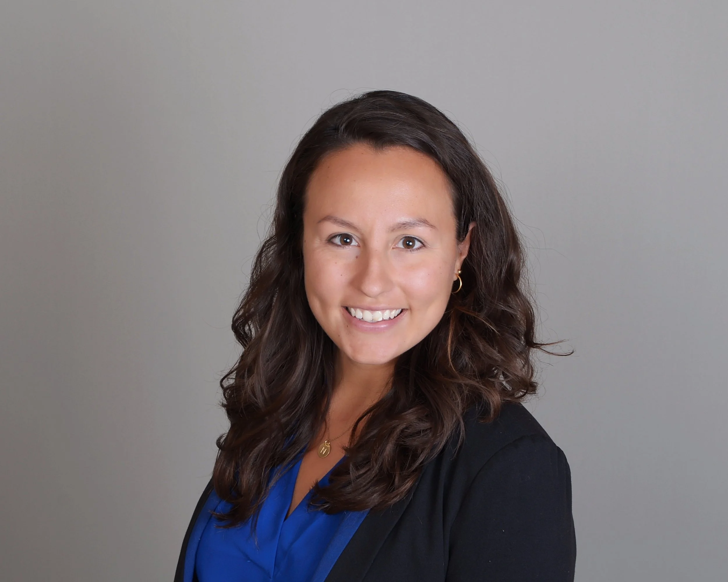A woman with brown, wavy hair, smiling, wearing gold hoop earrings and a black blazer over a blue top, against a plain light gray background.