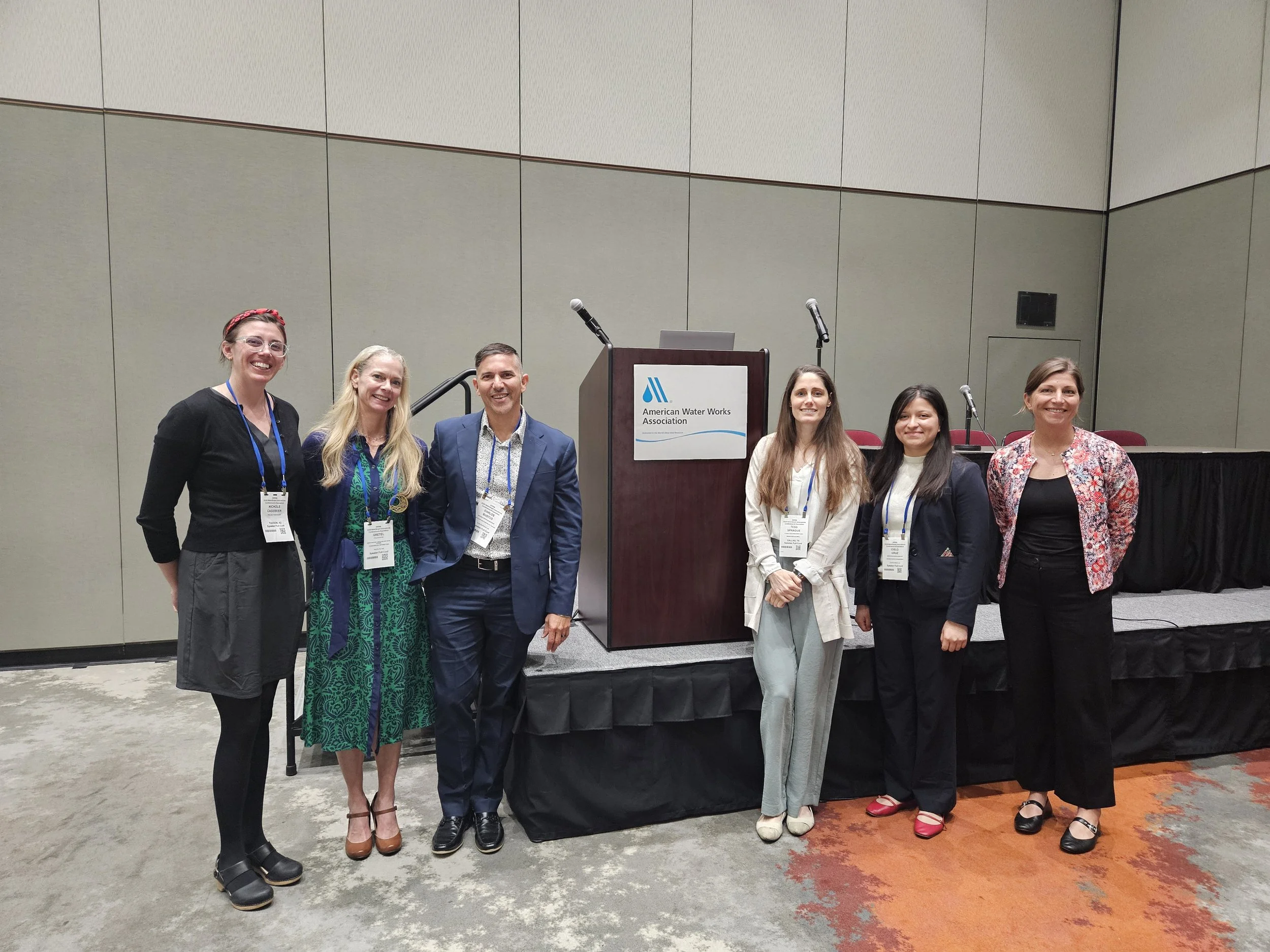 Group of six people standing in front of a podium with a sign that reads 'American Water Works Association' at a conference.