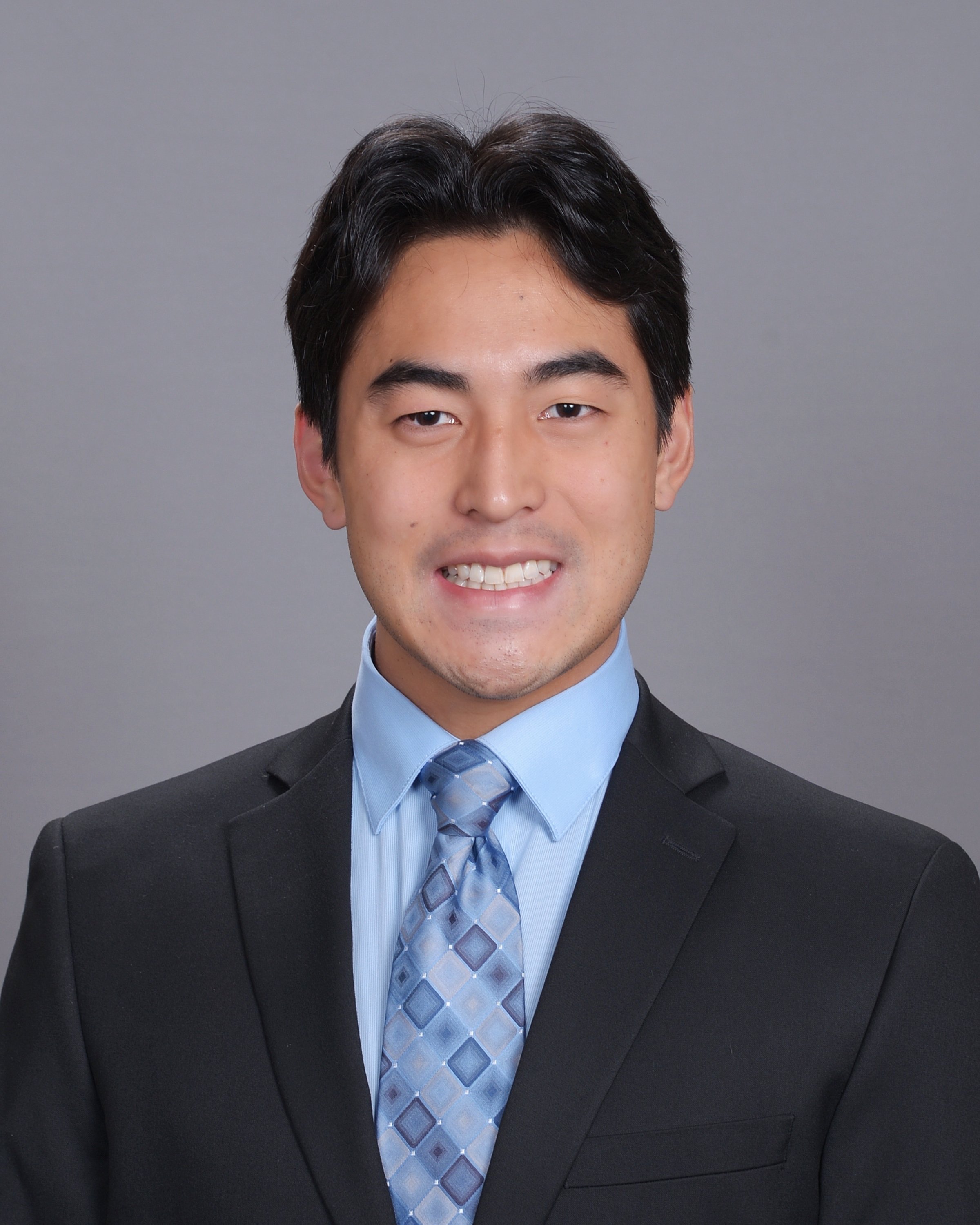Close-up of a smiling man in a suit, wearing a blue dress shirt and a patterned tie, against a plain gray background.