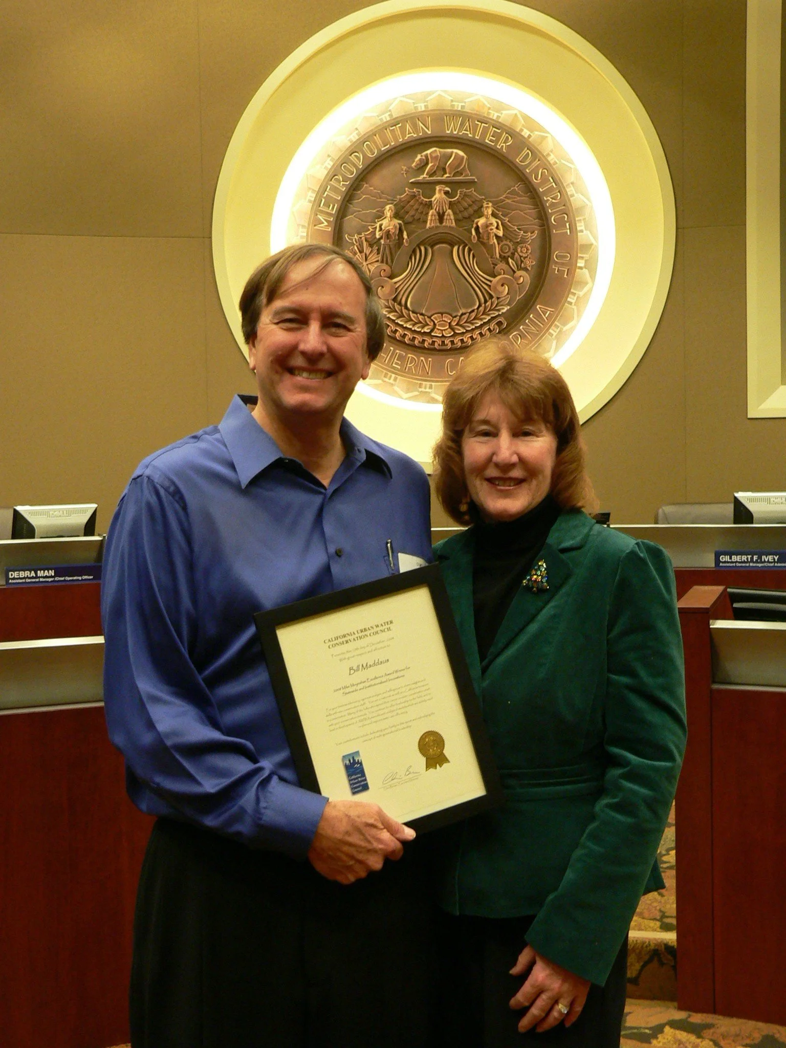 Two people standing in front of a large sign for the Metropolitan Water District of Southern California. The man is wearing a blue shirt, and the woman is wearing a dark green blazer with a decorative pin, holding a framed certificate. They are smiling and seem to be celebrating an award or recognition.