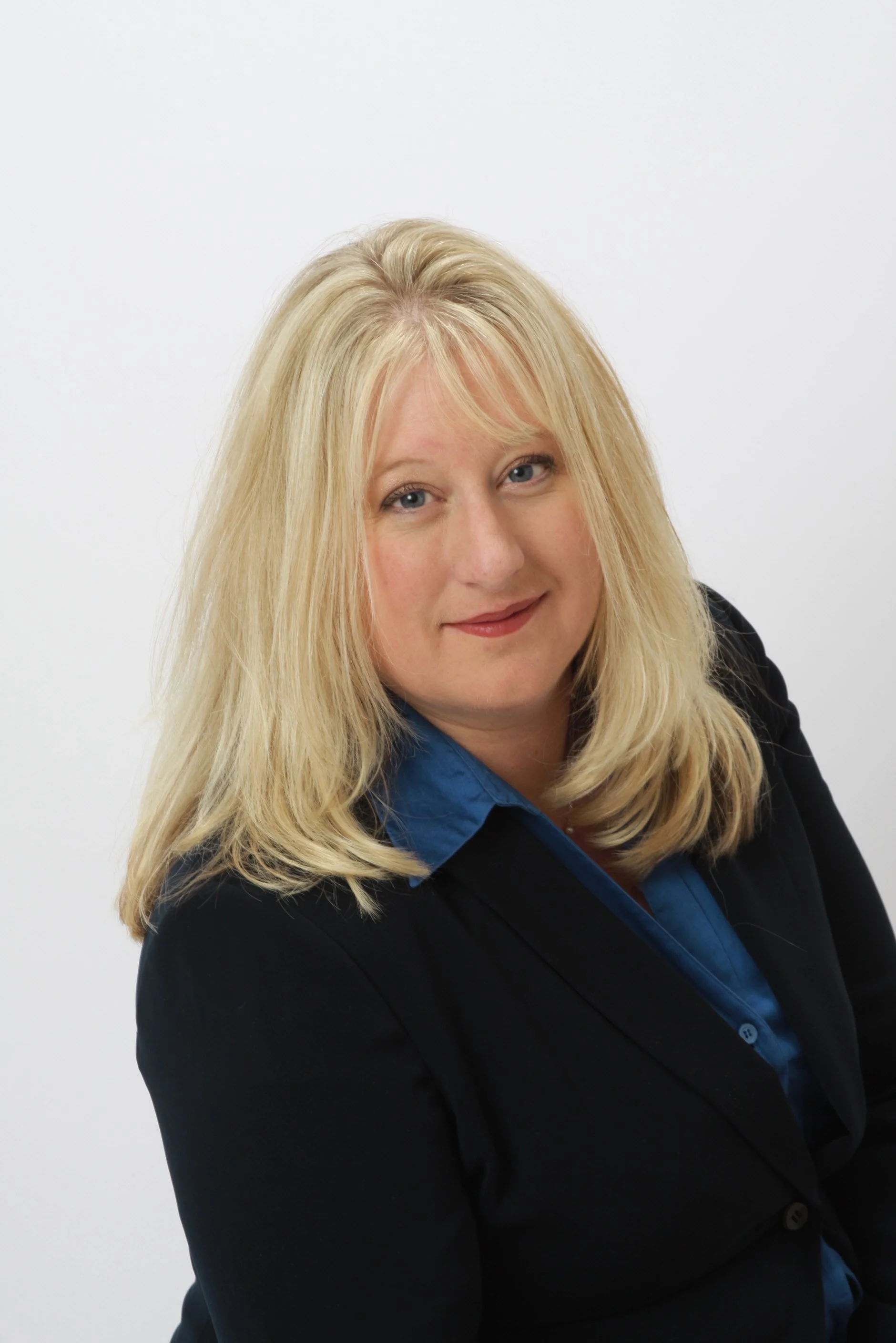 A woman with long blonde hair, blue eyes, and a friendly expression, wearing a black blazer and a blue blouse, posing against a plain white background.