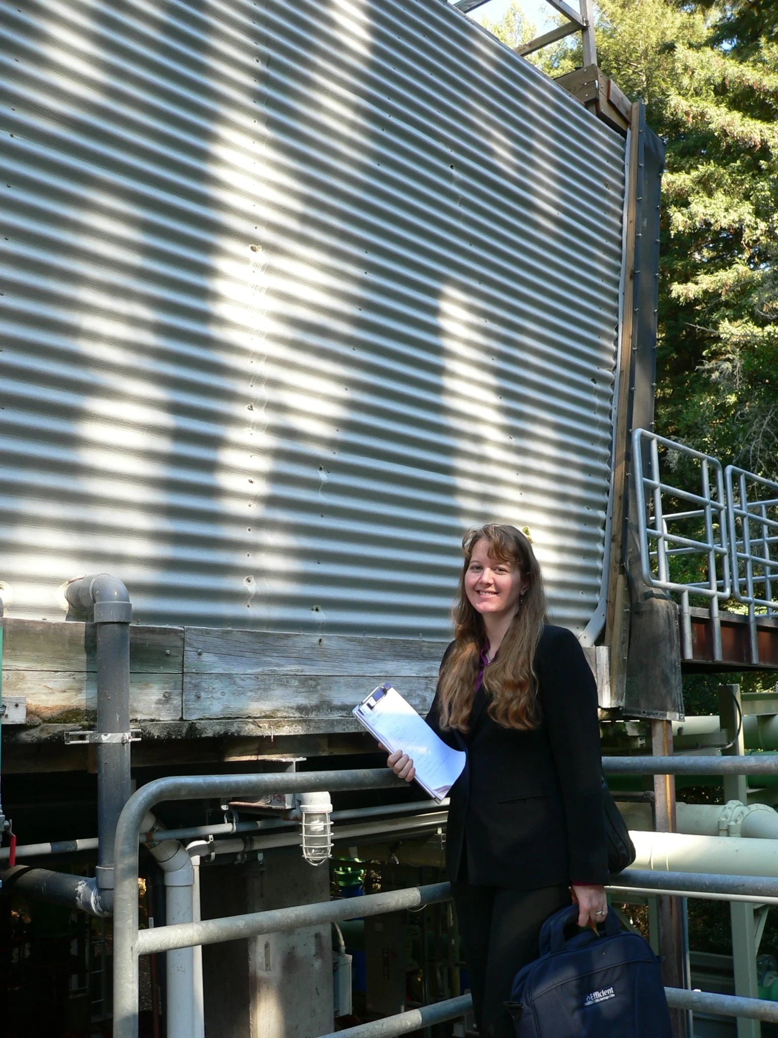 A young woman with long brown hair holding a clipboard and wearing a black jacket and pants, standing outdoors near a large industrial metal tank and pipes, with trees in the background.