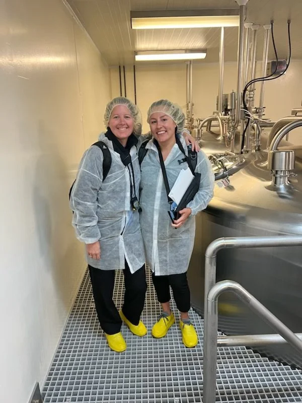Two women in protective clothing, hairnets, and yellow shoe covers standing inside a biotech or pharmaceutical manufacturing facility, smiling at the camera.
