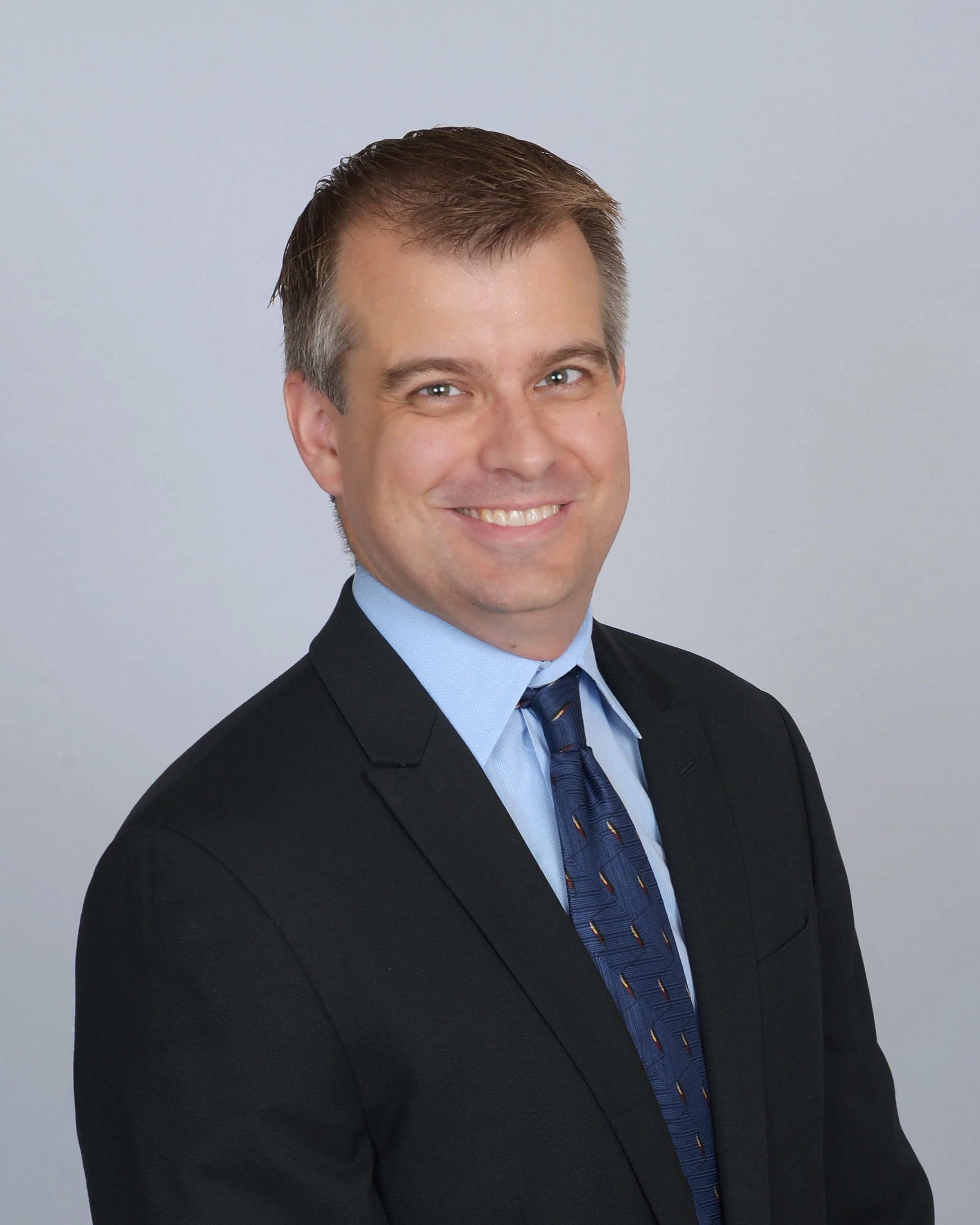 Headshot of a smiling man with short brown hair, wearing a dark suit, light blue shirt, and a blue tie with yellow and red stripe patterns, against a light gray background.