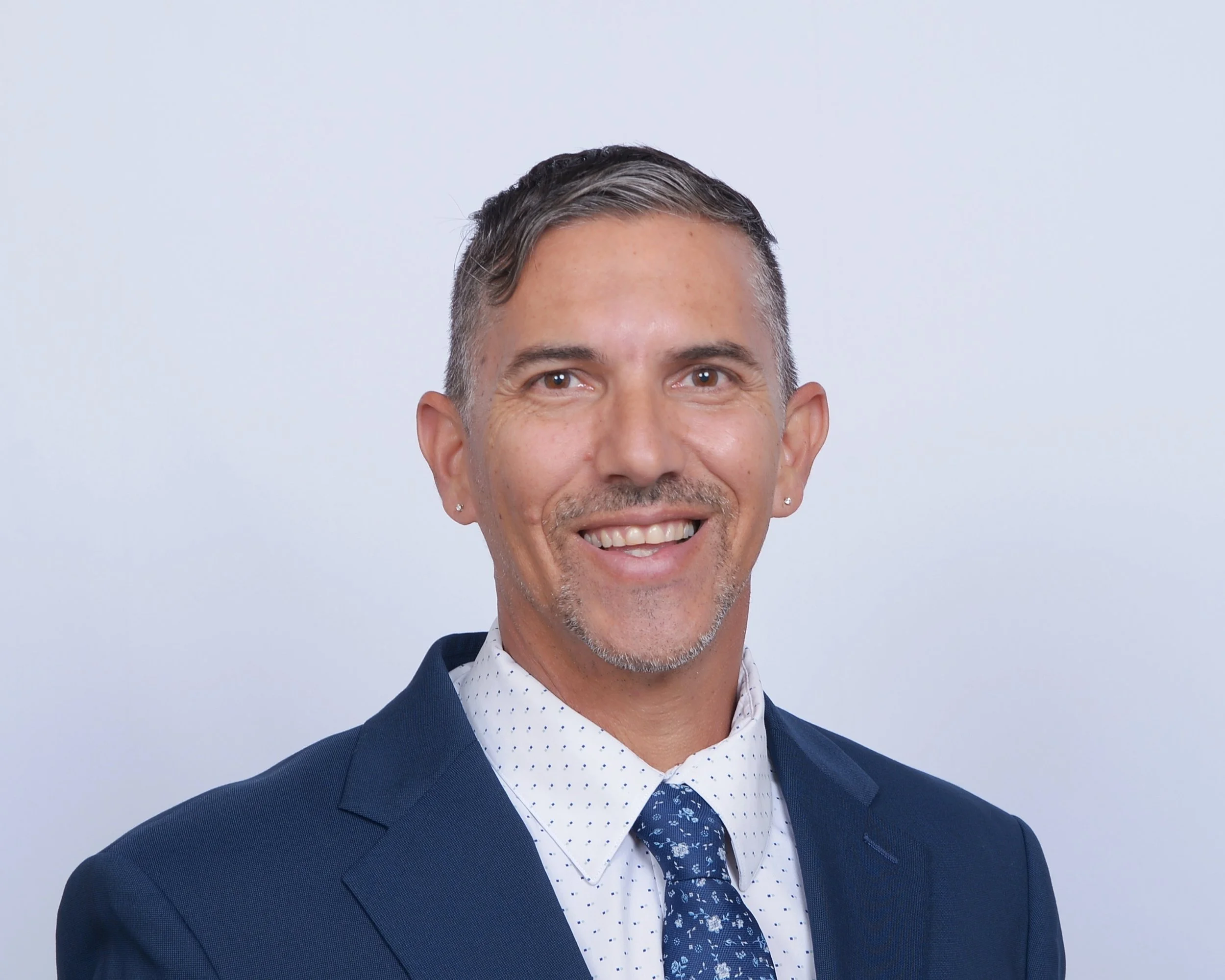 Close-up of a smiling man with gray hair in a side part, wearing a navy blue suit, white shirt with small polka dots, and a navy floral tie, against a plain light background.