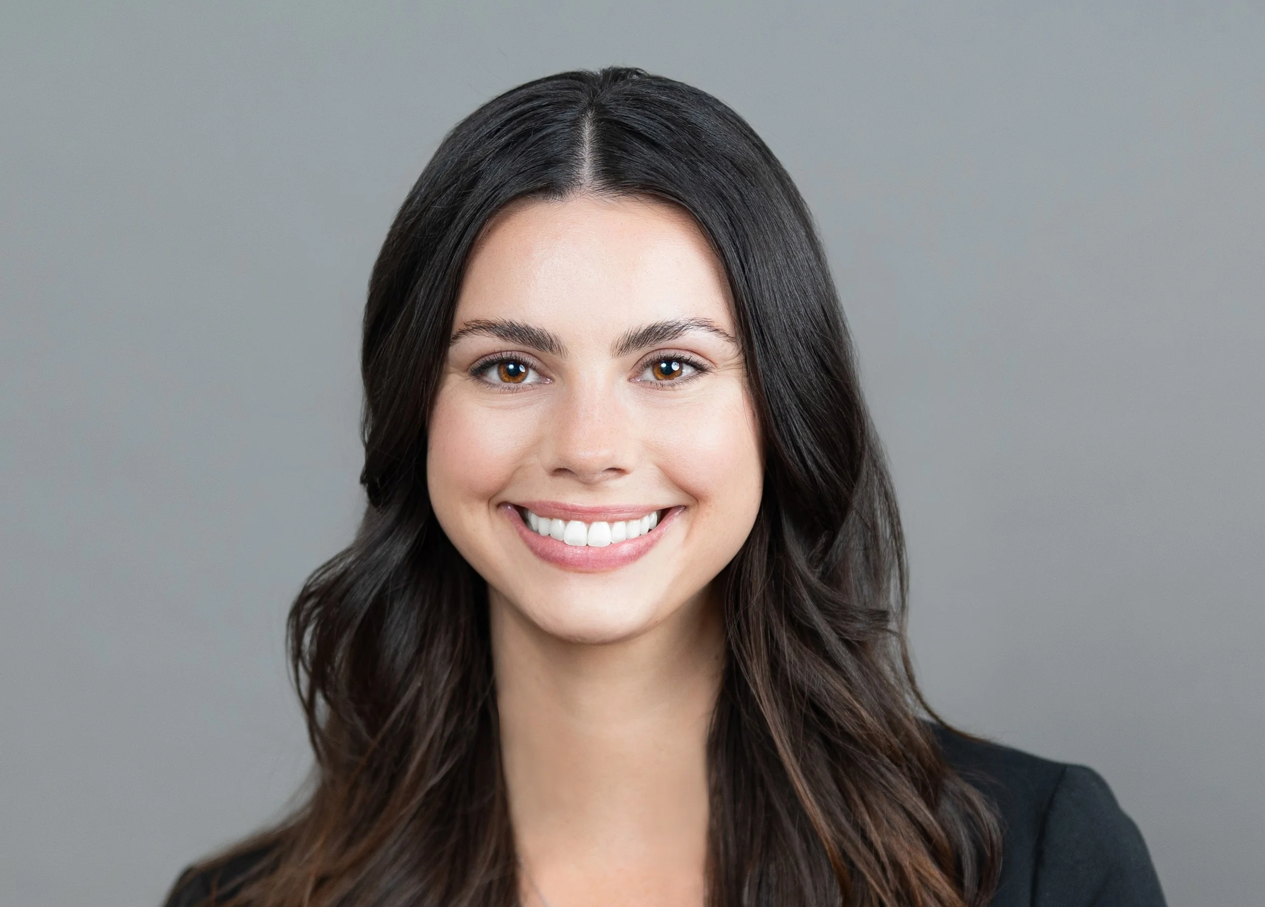 Close-up of a smiling woman with long brown hair and brown eyes, wearing a dark blazer, against a neutral gray background.
