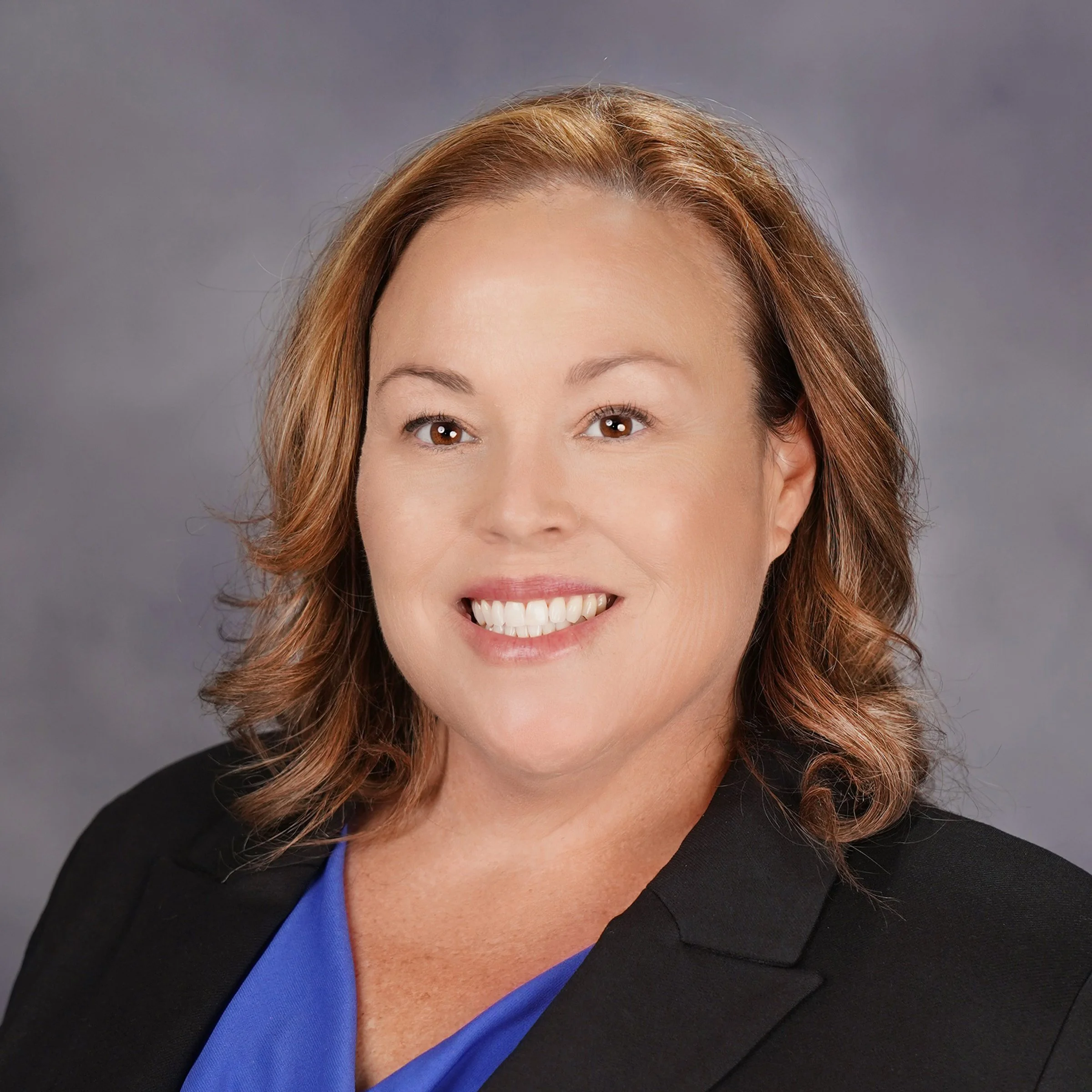 Close-up of an Asian woman's face, showing her smiling with white teeth, light skin, brown eyes, and reddish-brown wavy hair. She is wearing a black blazer and a blue top, with a gray background.