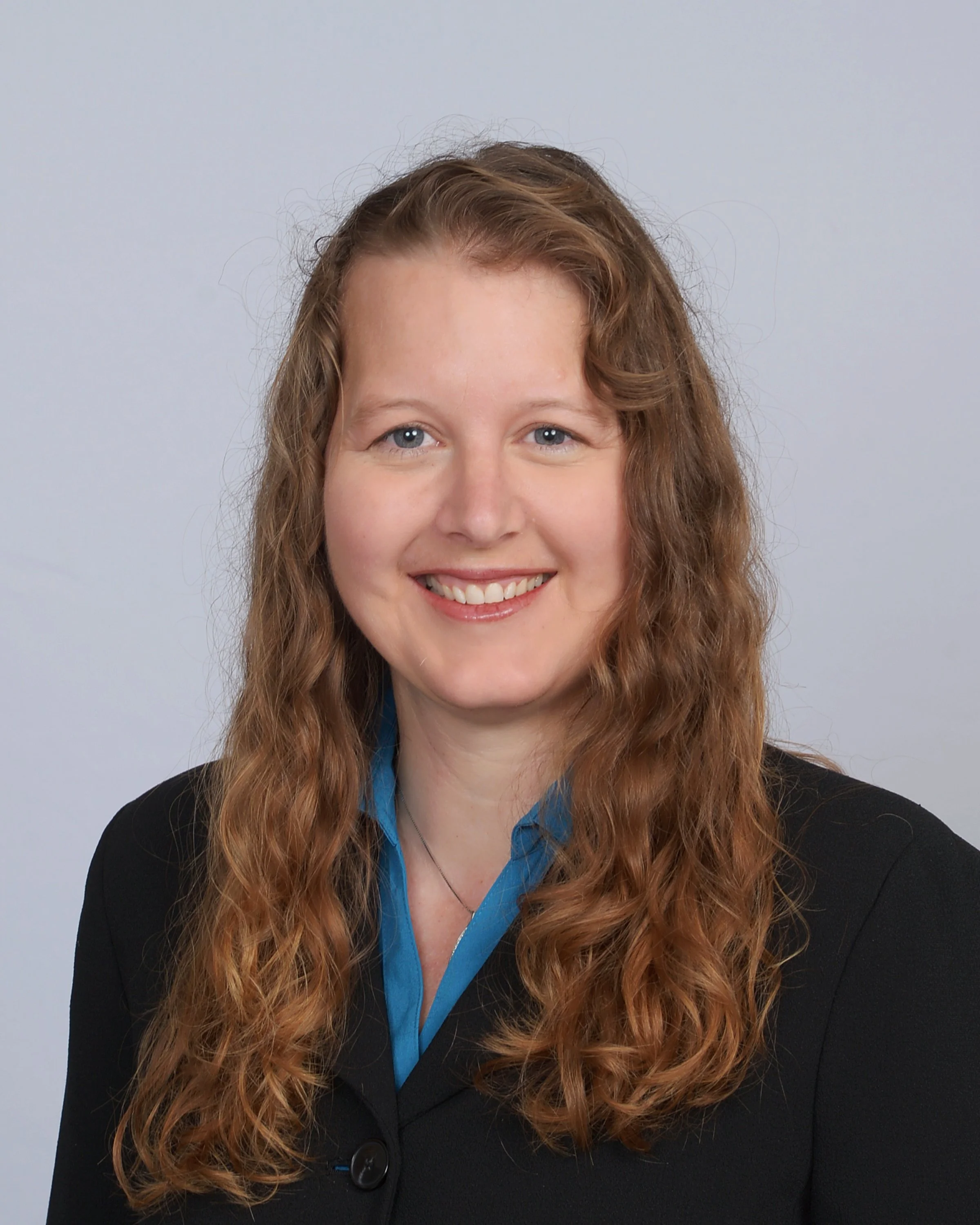Portrait of a smiling woman with long, wavy red hair, wearing a black blazer over a blue shirt, standing against a plain light background.
