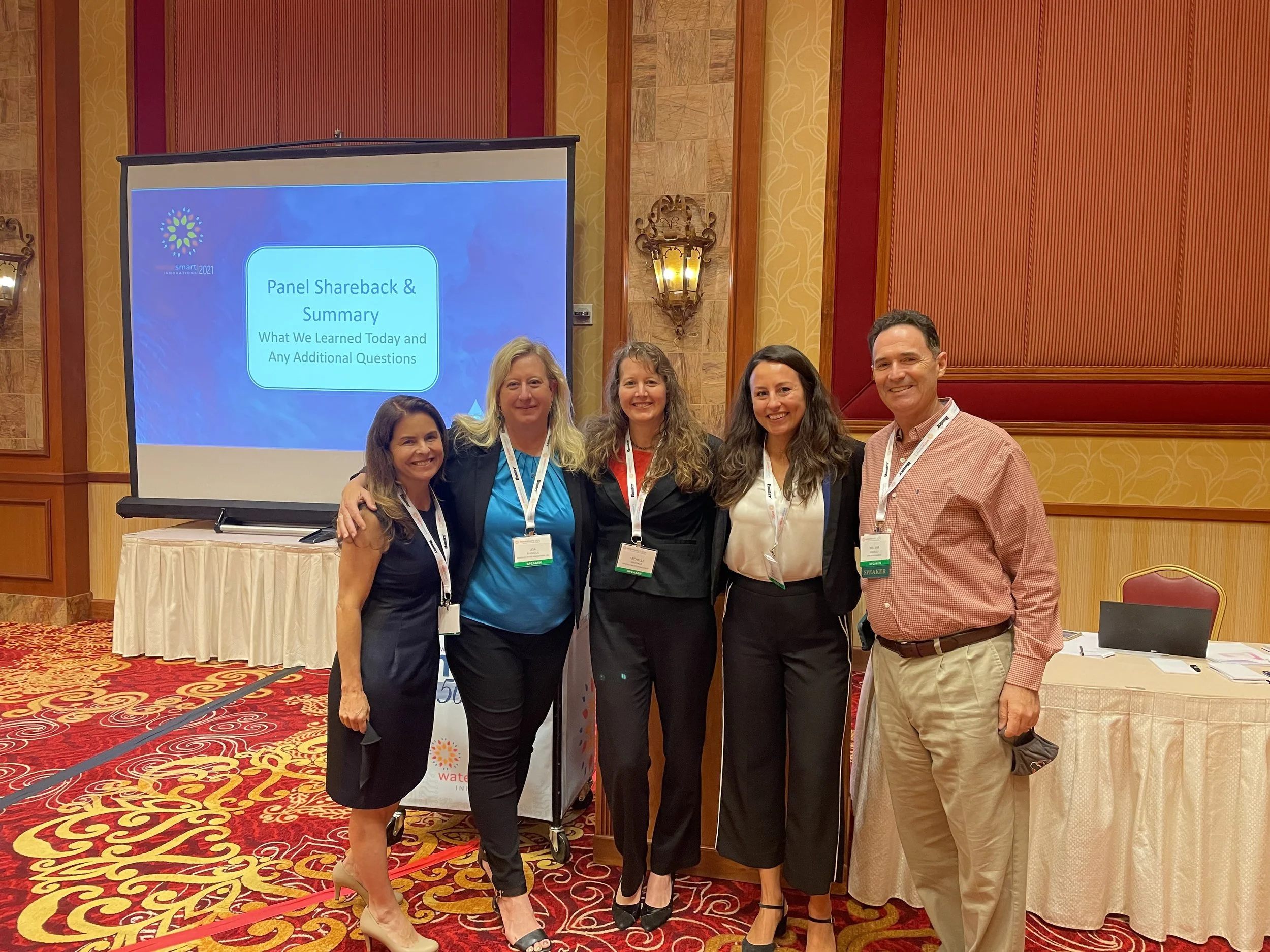 A group of five people standing together in front of a presentation screen at a conference or event. The screen displays the title 'Panel Shareback & Summary' and a subtitle. The background consists of ornate red, gold, and wood-paneled walls, and the floor has a patterned red carpet. The people are smiling and wearing conference name badges.