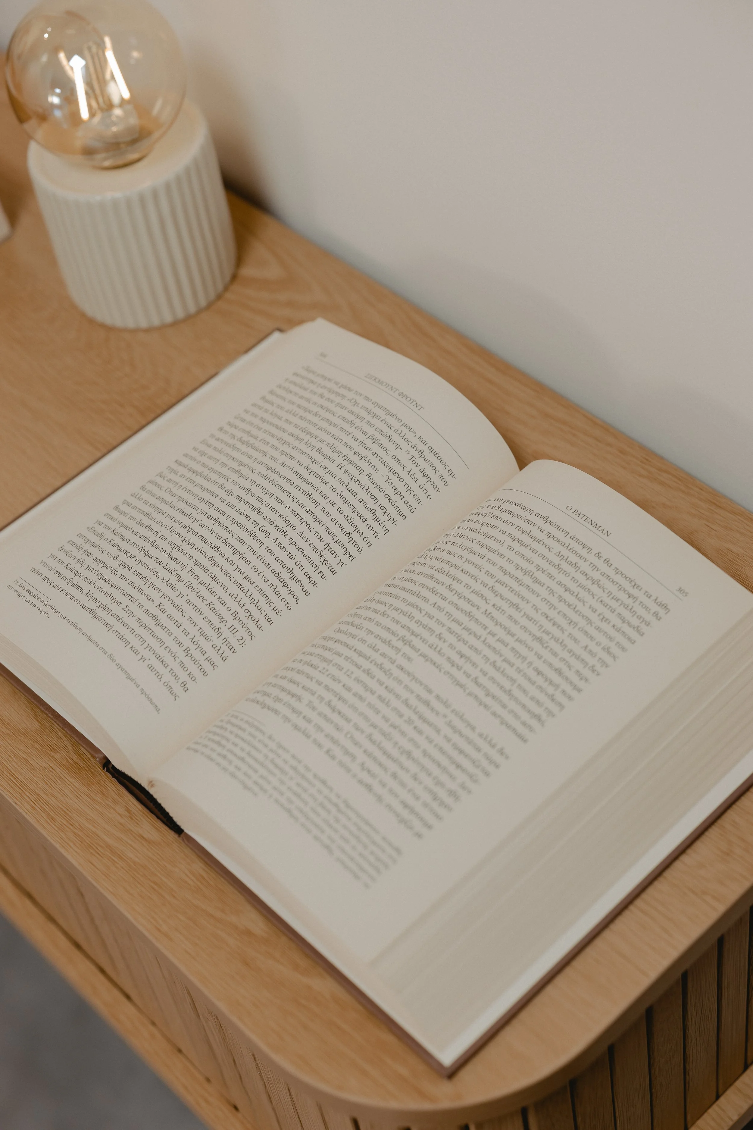 An open book on a wooden desk next to a modern table lamp with a ribbed ceramic base and a large clear lightbulb.