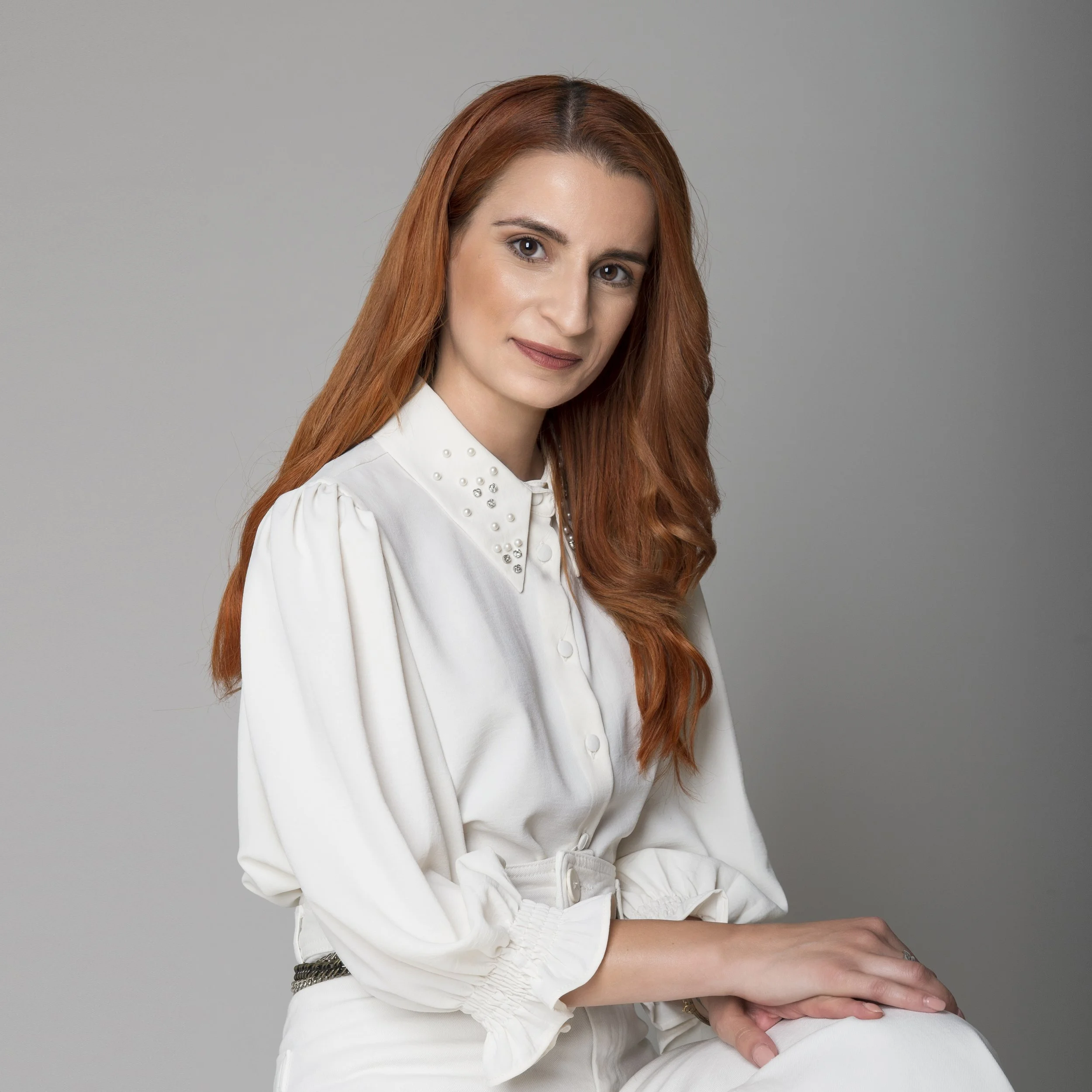 A woman with long red hair wearing a white blouse with pearl embellishments on the collar, sitting against a plain gray background.