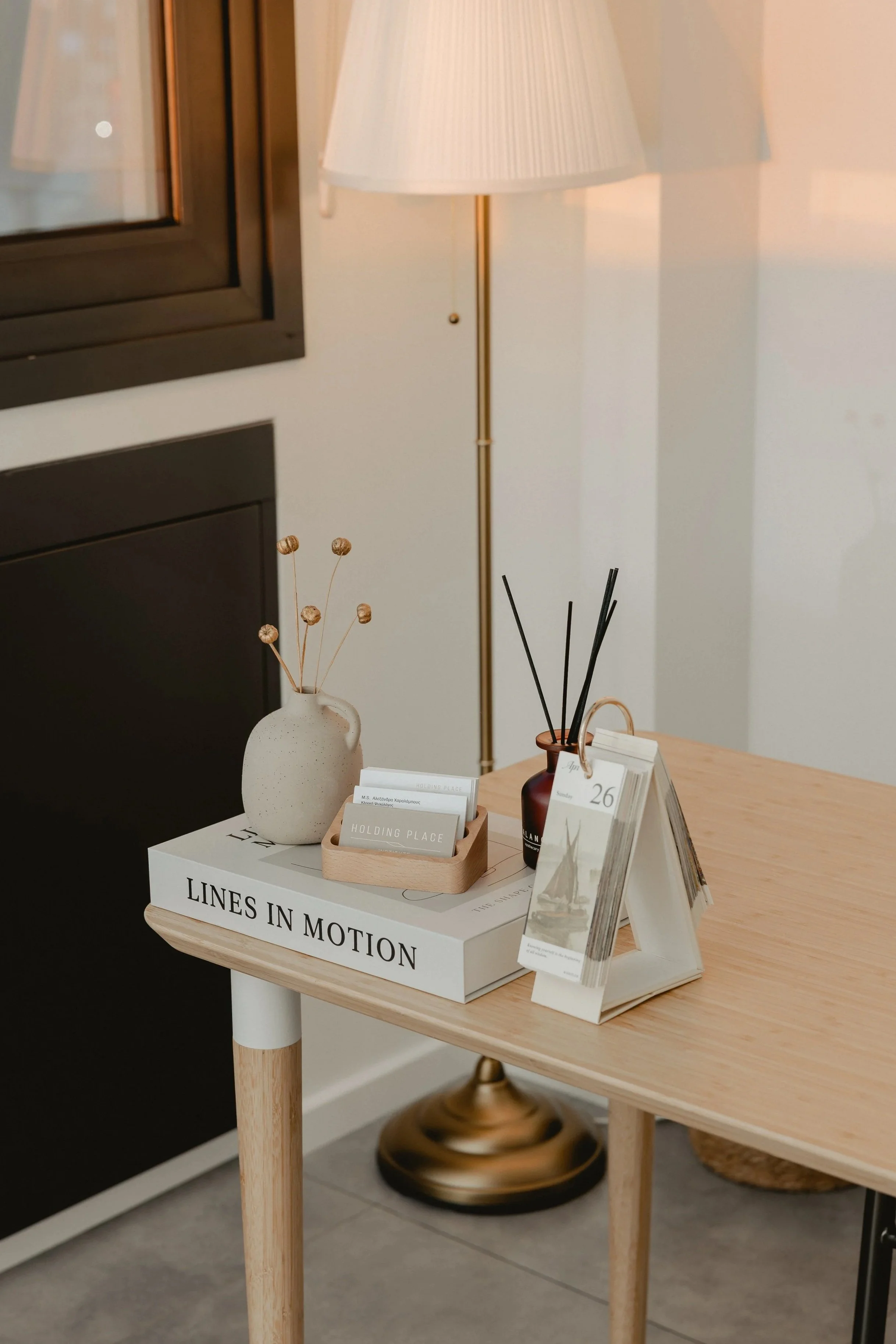 A wooden side table with a white book titled 'LINES IN MOTION' on top. The table has a white vase with dried flower stems, a small wooden tray with cards or coasters, a dark red container holding black reed diffusers, and brochures on a white display stand. In the background, there is a floor lamp with a white shade, a window with a black frame, and a black and gold decorative object underneath.
