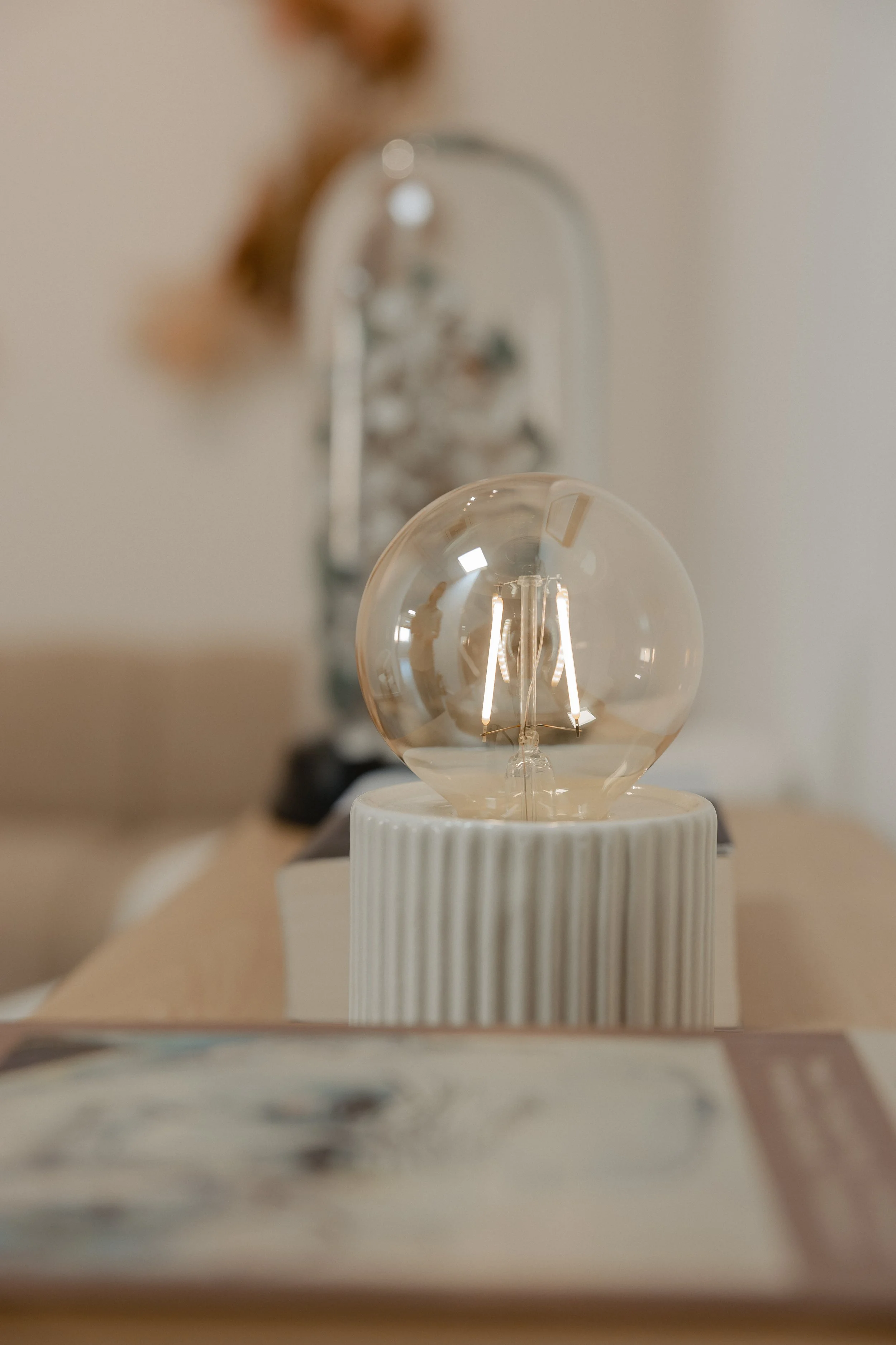Close-up of an illuminated Edison-style light bulb on a striped white base, with a blurred background featuring a metallic faucet and a wall.