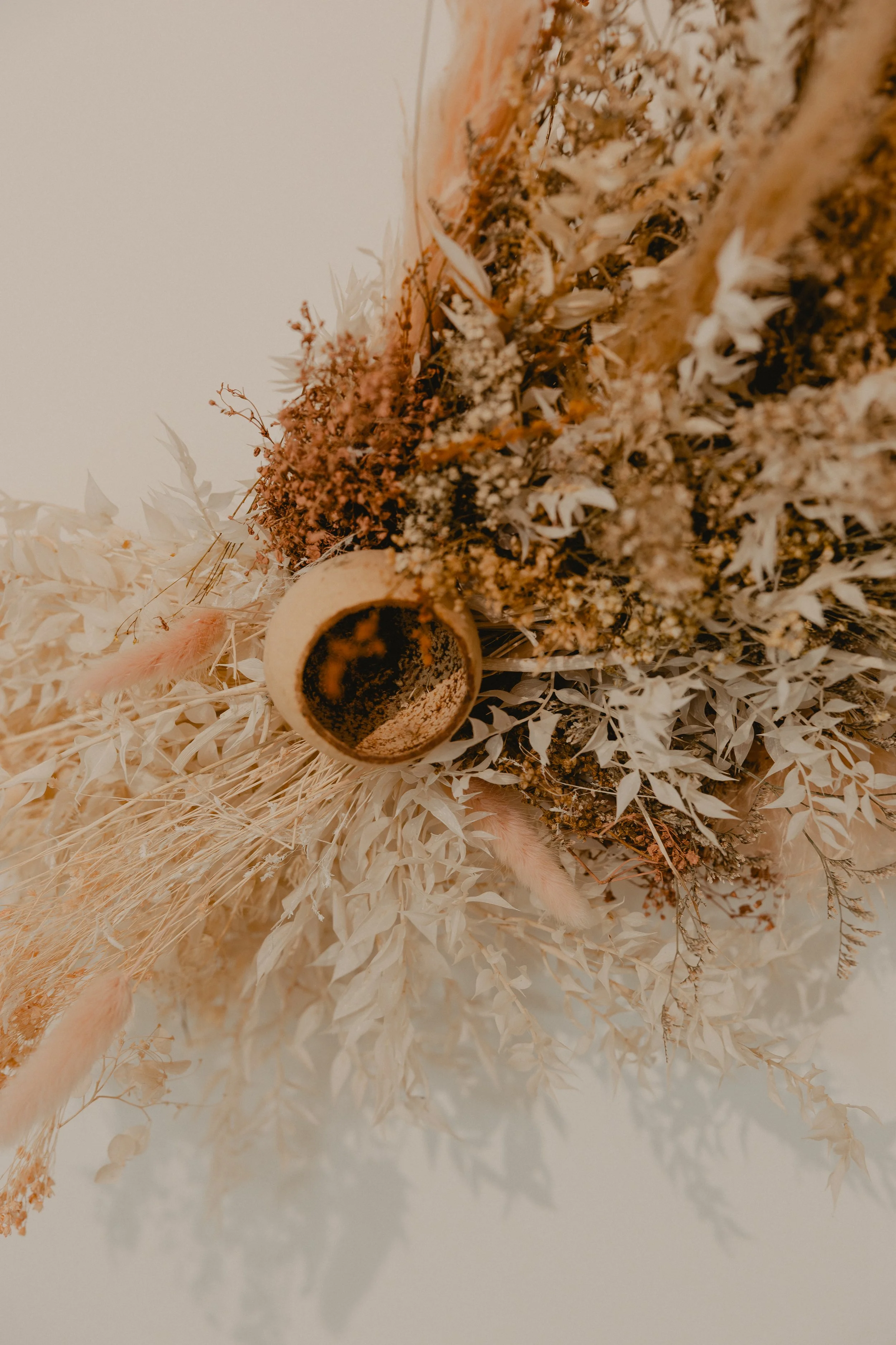 A collection of dried flowers and herbs, including white, pink, and beige-colored foliage, arranged artfully with a small, textured cup or container among the dried botanicals.