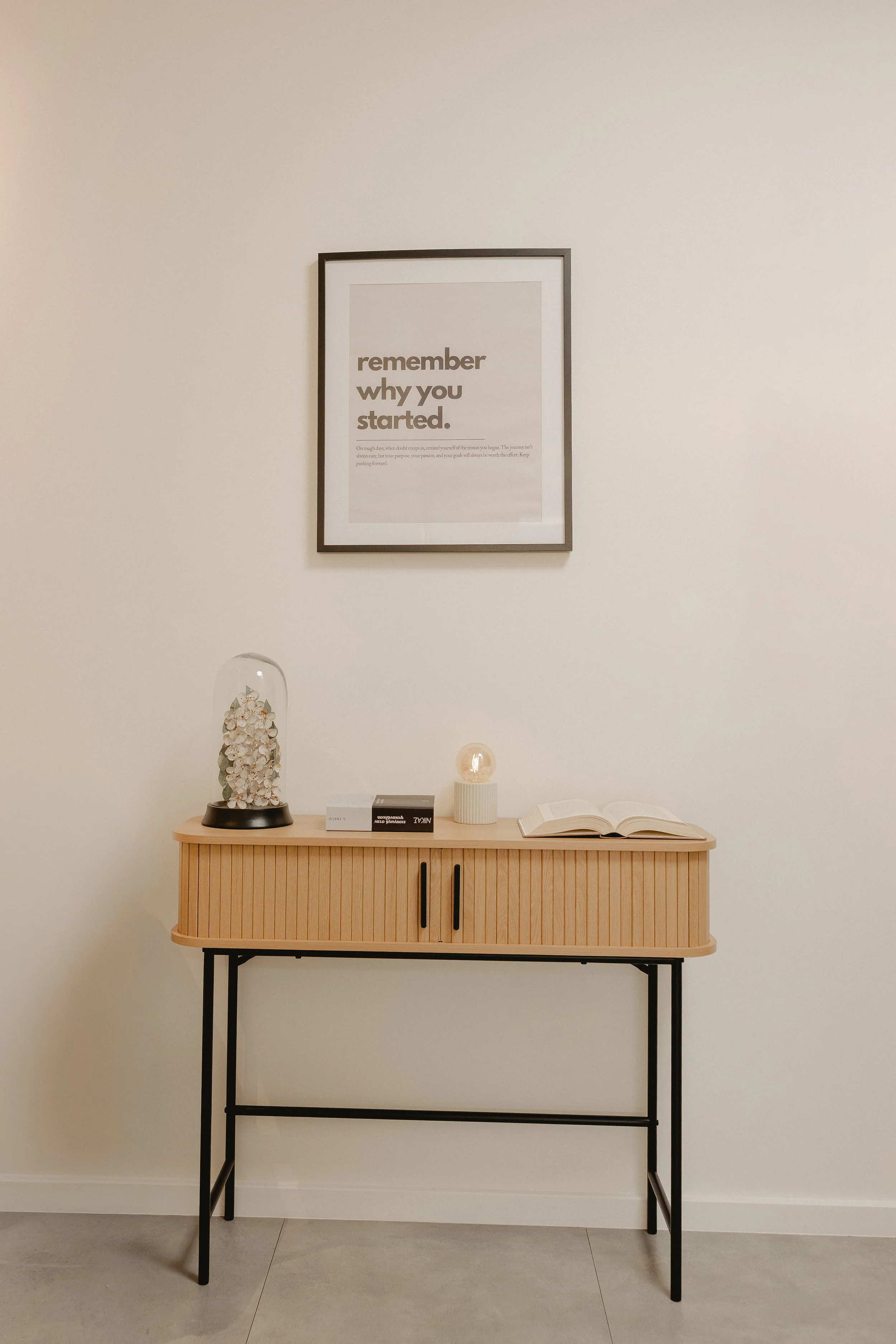 A minimalist wooden sideboard with black metal legs against a plain white wall. On top are decorative items, including a glass container filled with white shells, three books, a small white lamp with a glowing bulb, and an open book. Above the sideboard hangs a framed quote that reads, 'remember why you started.'