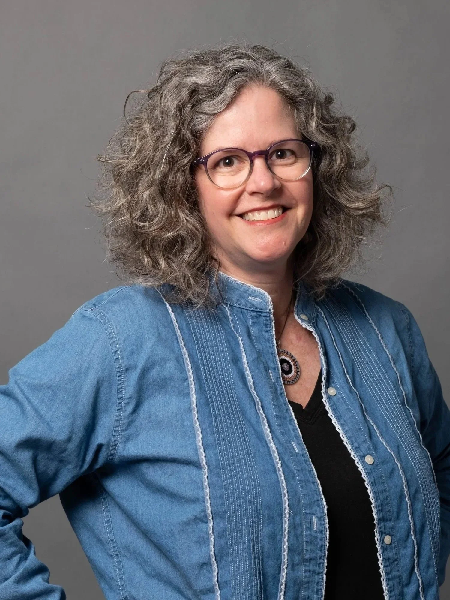Portrait of a smiling woman with shoulder length curly silvering hair with round purple glasses and a denim shirt.