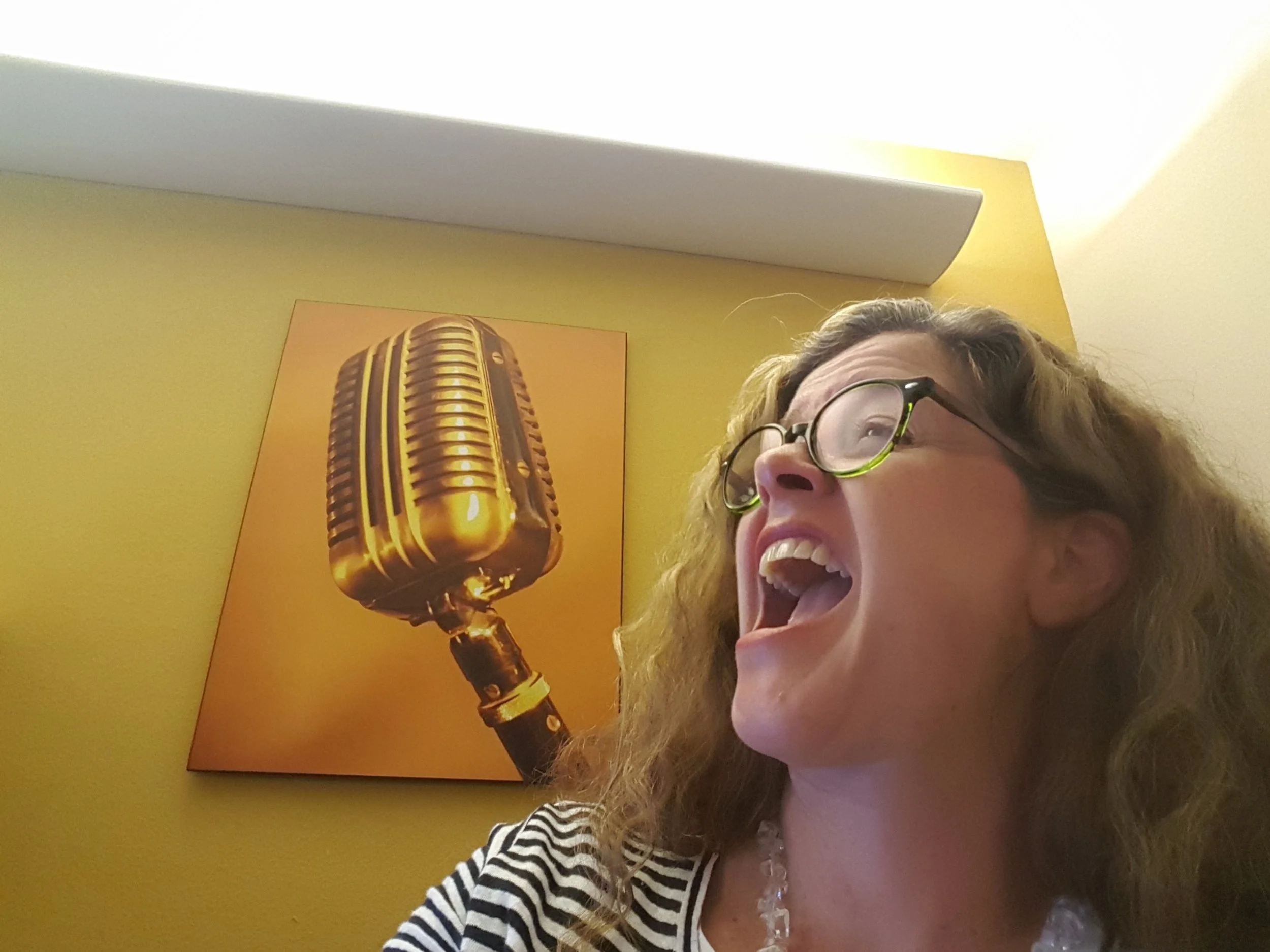 Kerry, mouth wide in song in front of large wall art of a microphone.