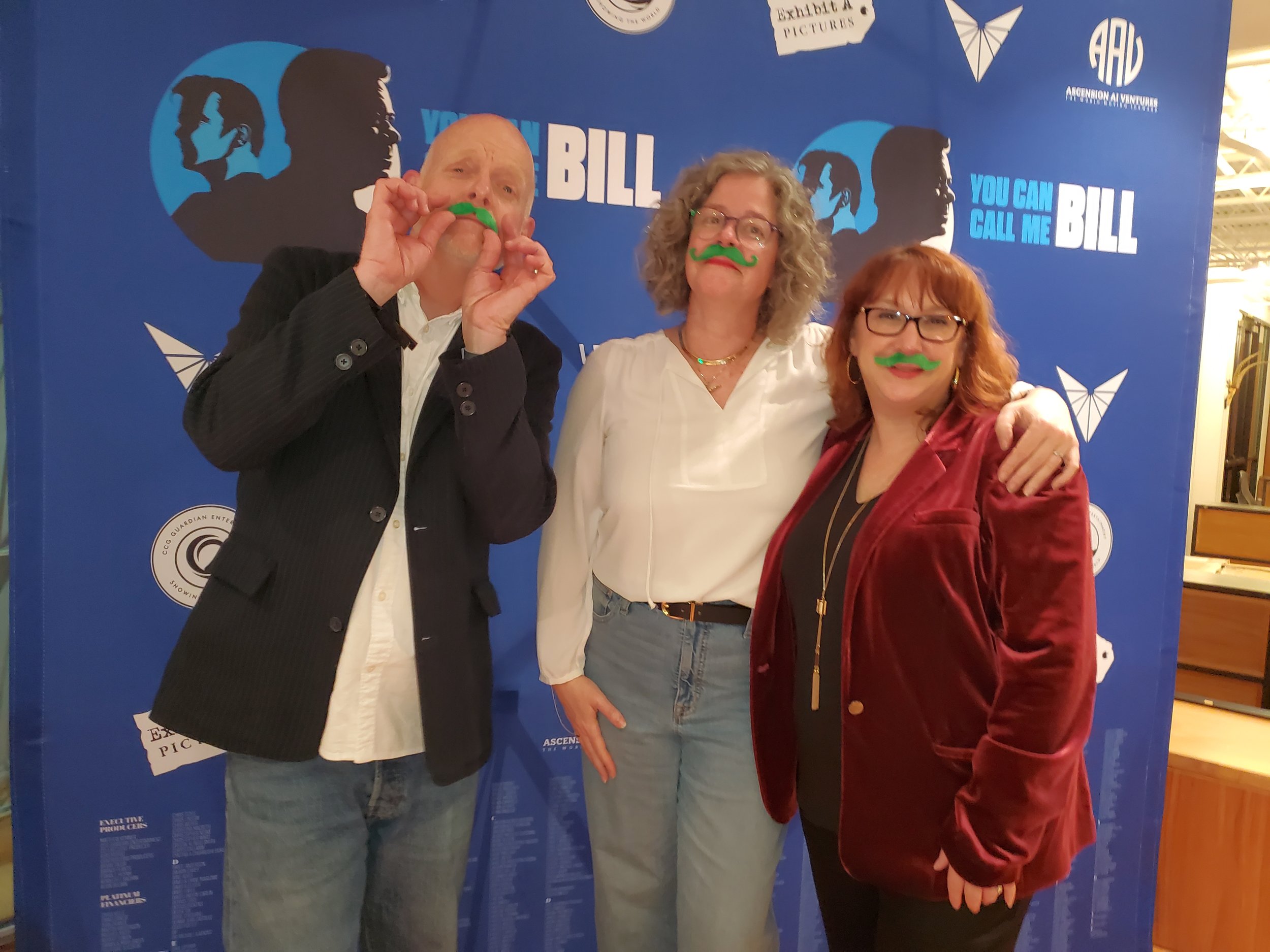 Kerry between two colleagues at a premiere step and repeat wearing silly green mustaches.