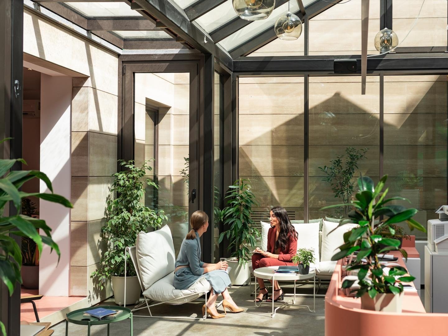 Two women sitting and talking in a glass-enclosed indoor space with plants, modern furniture, and natural lighting.