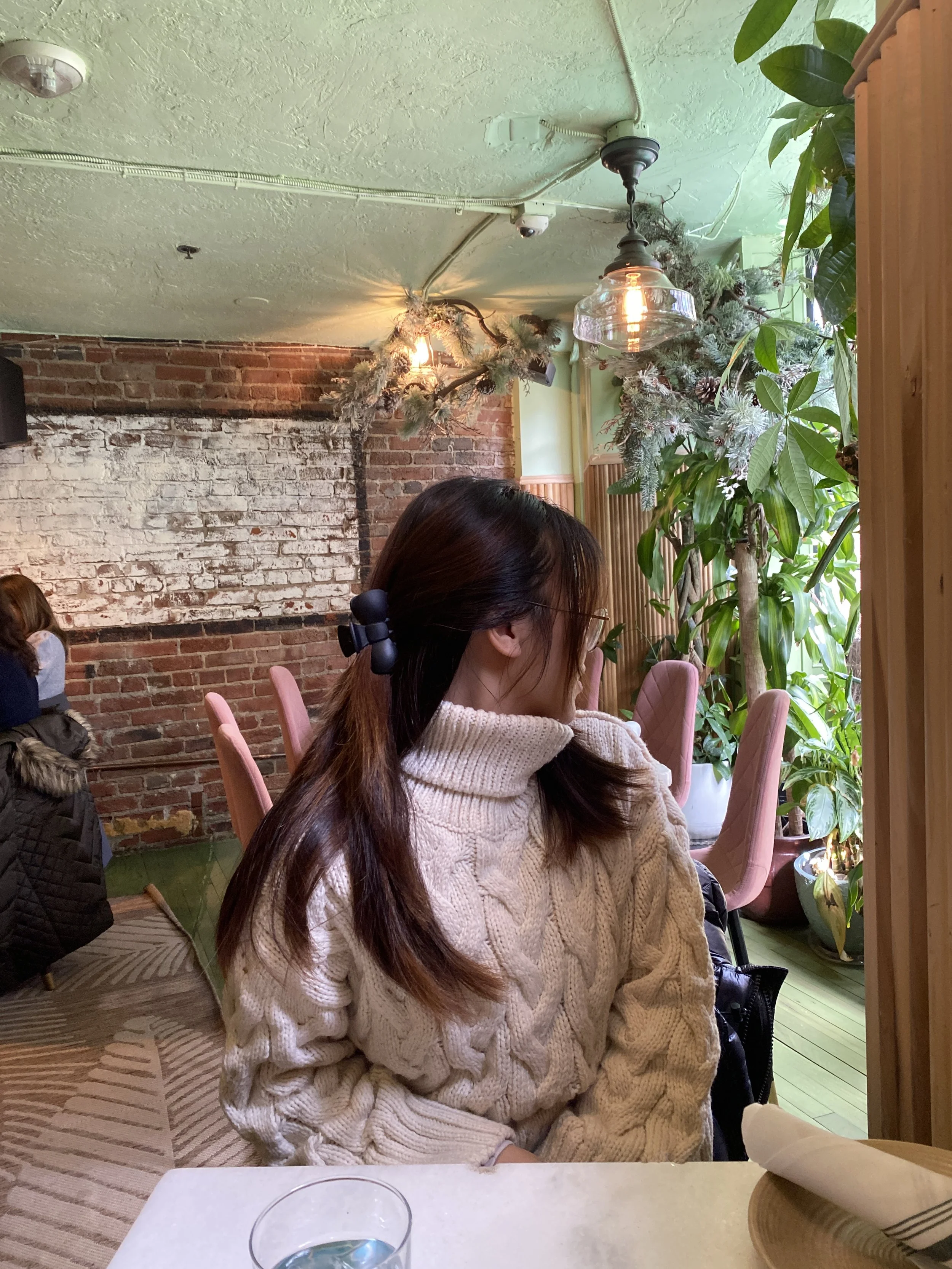 A woman with brown hair tied back, wearing a chunky cream-colored cable knit sweater, sitting at a table in a cozy restaurant with exposed brick wall and large green plants. She appears to be looking out the window or at something outside.