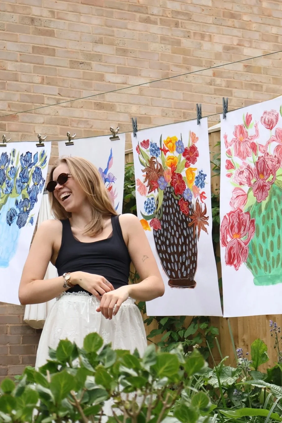 A woman wearing sunglasses, a black tank top, and white skirt standing outdoors in front of colorful floral paintings hanging on a clothesline wall. She is smiling and appears happy.