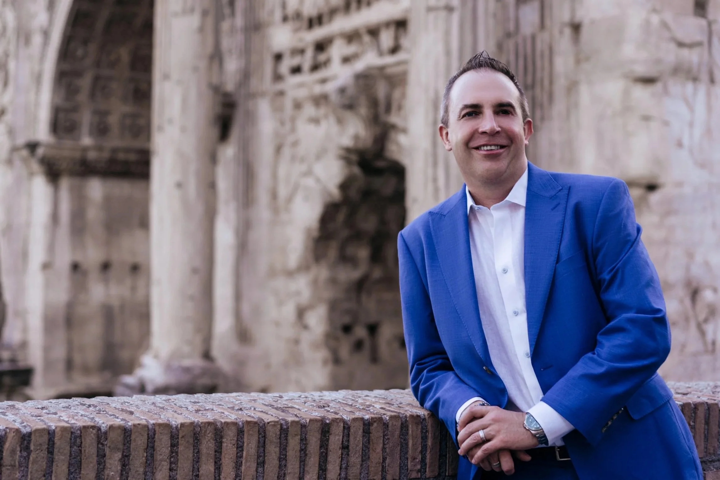 A man in a blue suit smiling and posing outdoors in front of ancient ruins or sculptures.