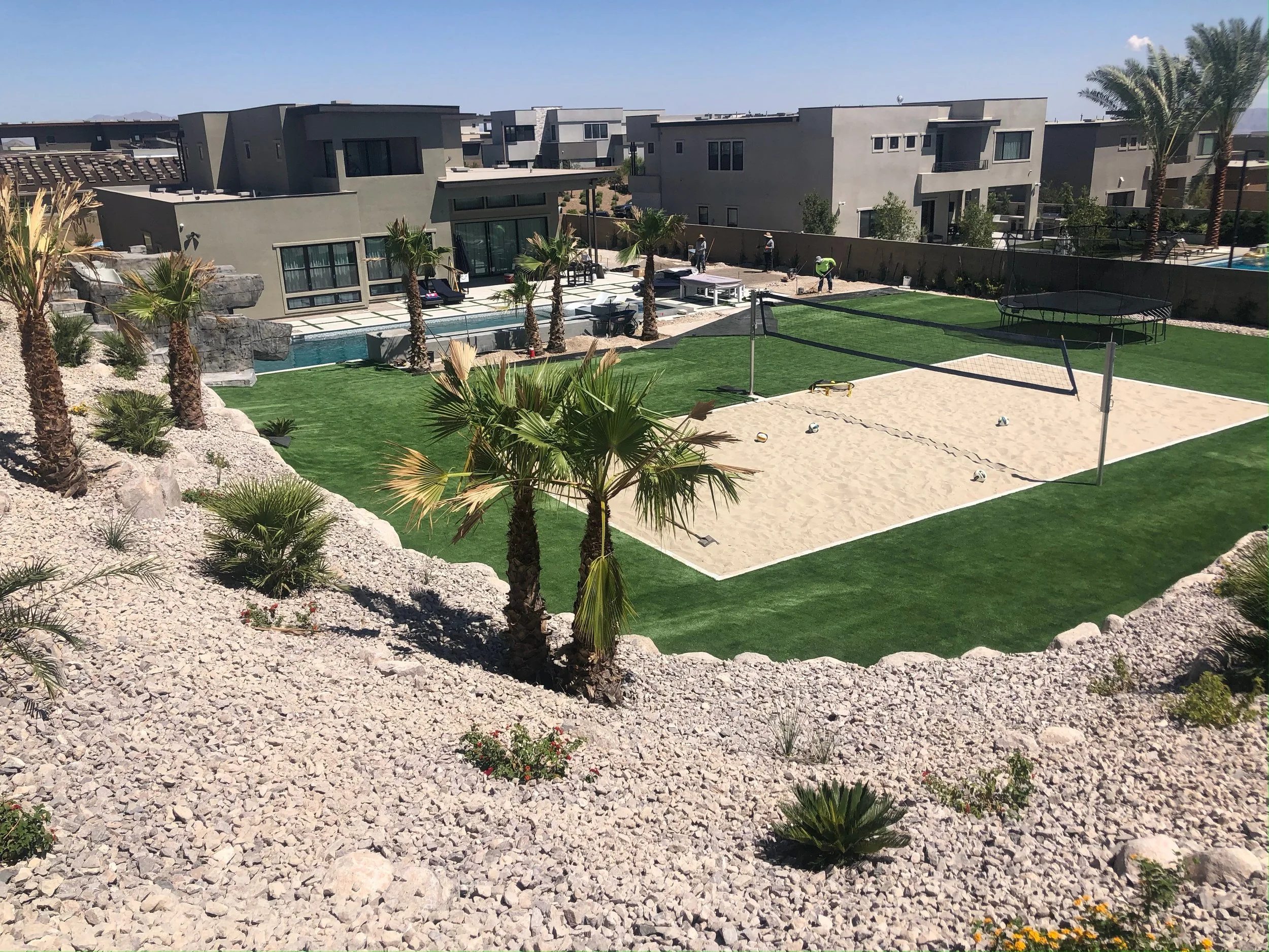A backyard with artificial grass, a volleyball net, a volleyball, trees, and a patio with chairs and a pool.