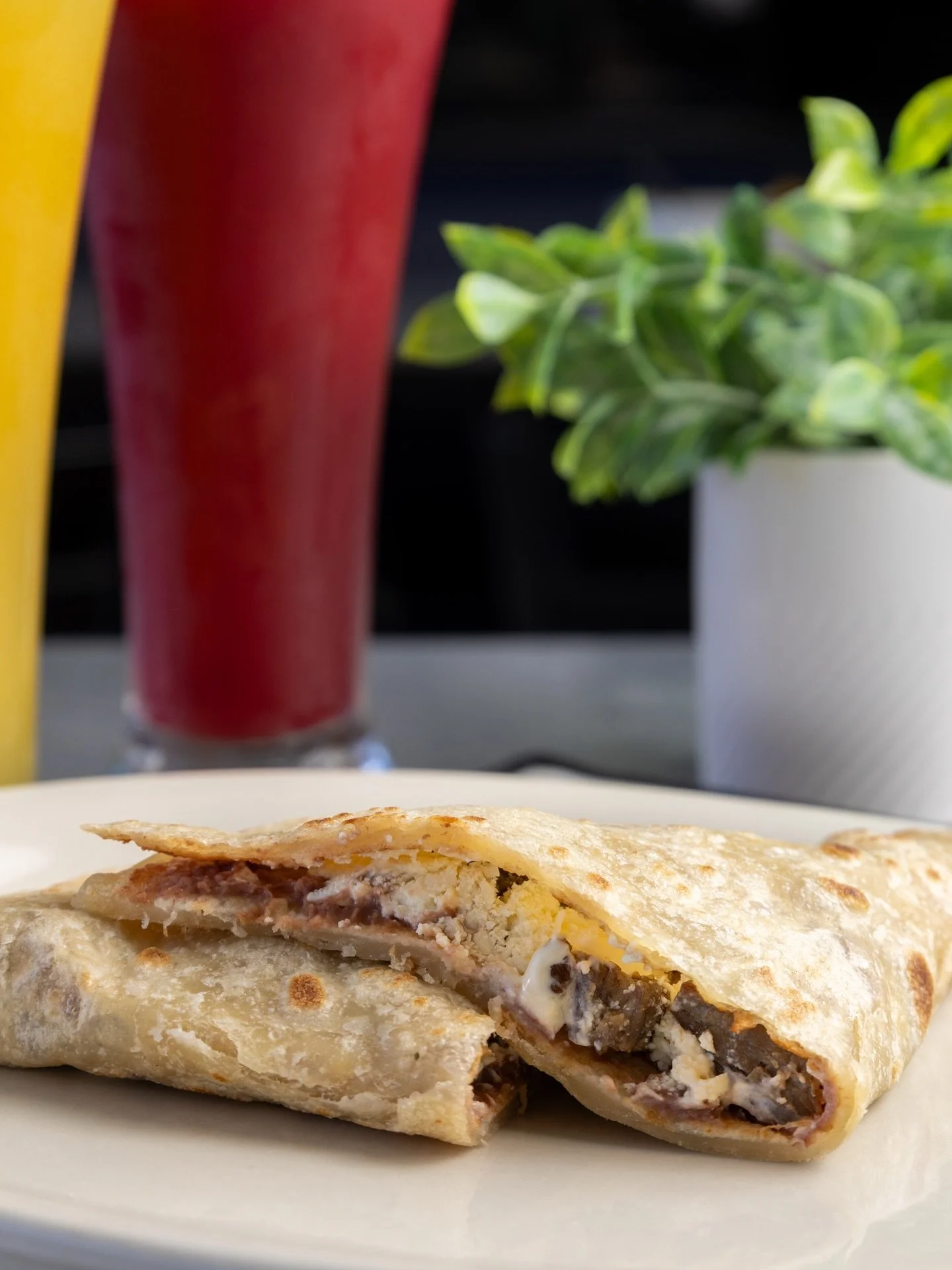 A quesadilla on a white plate with melted cheese and diced steak inside, accompanied by two drinks in the background and a potted plant to the side.