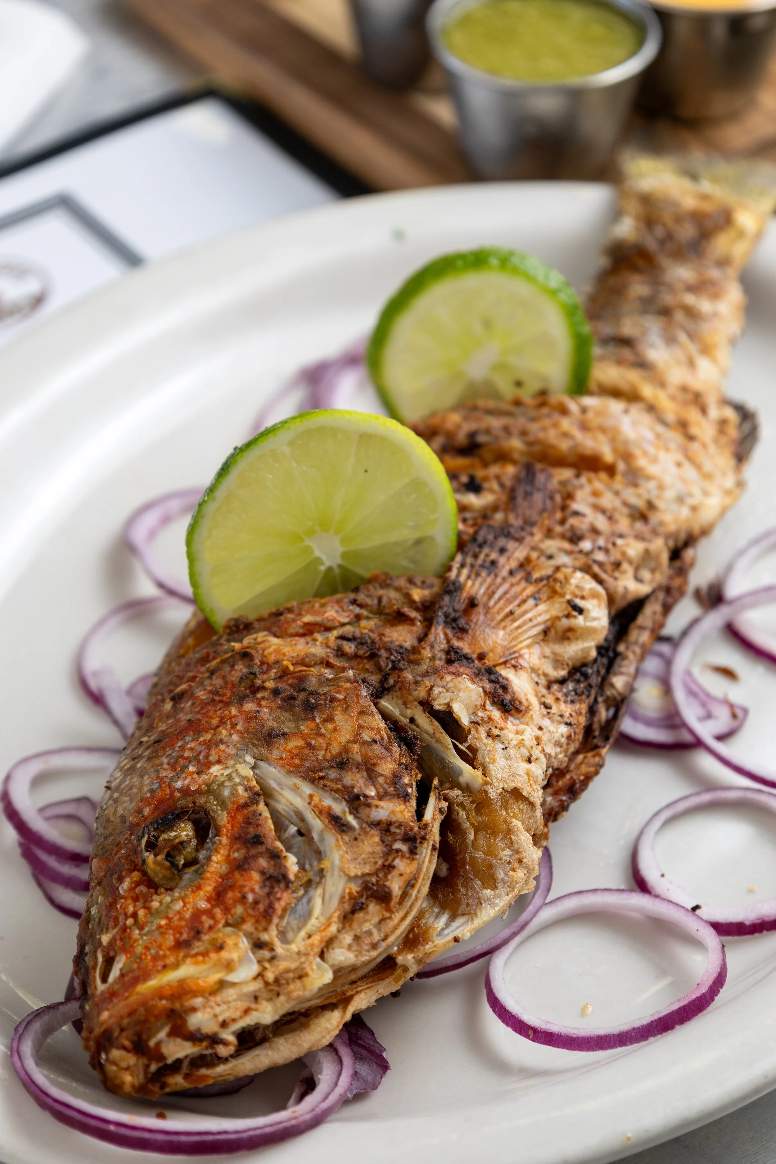 Whole grilled fish garnished with lime slices, surrounded by sliced red onions, served on a white plate with a small container of green sauce in the background.