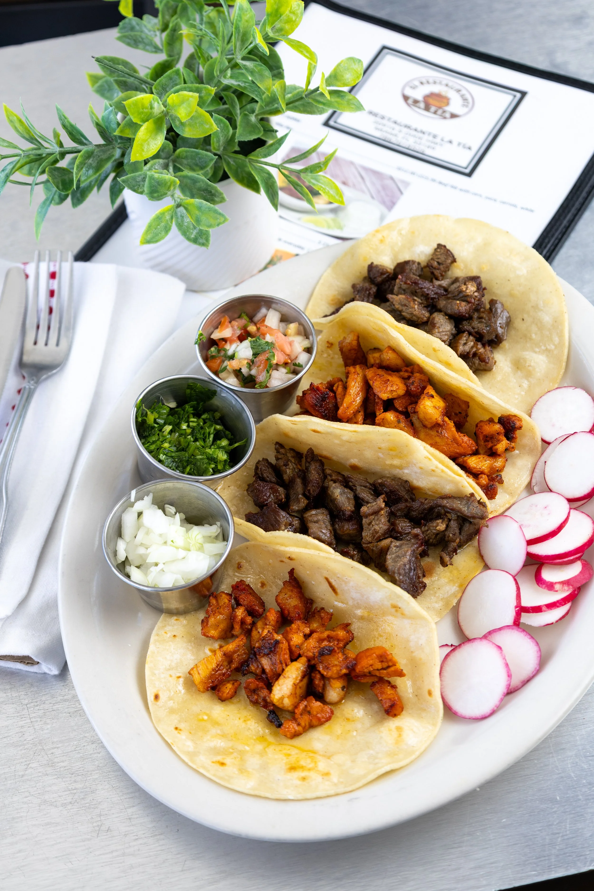 A plate of four street tacos with grilled beef, chicken, and pork, served with sliced radishes, diced onions, chopped cilantro, and salsas, on a white plate with a small potted plant, utensils, and a menu in the background.