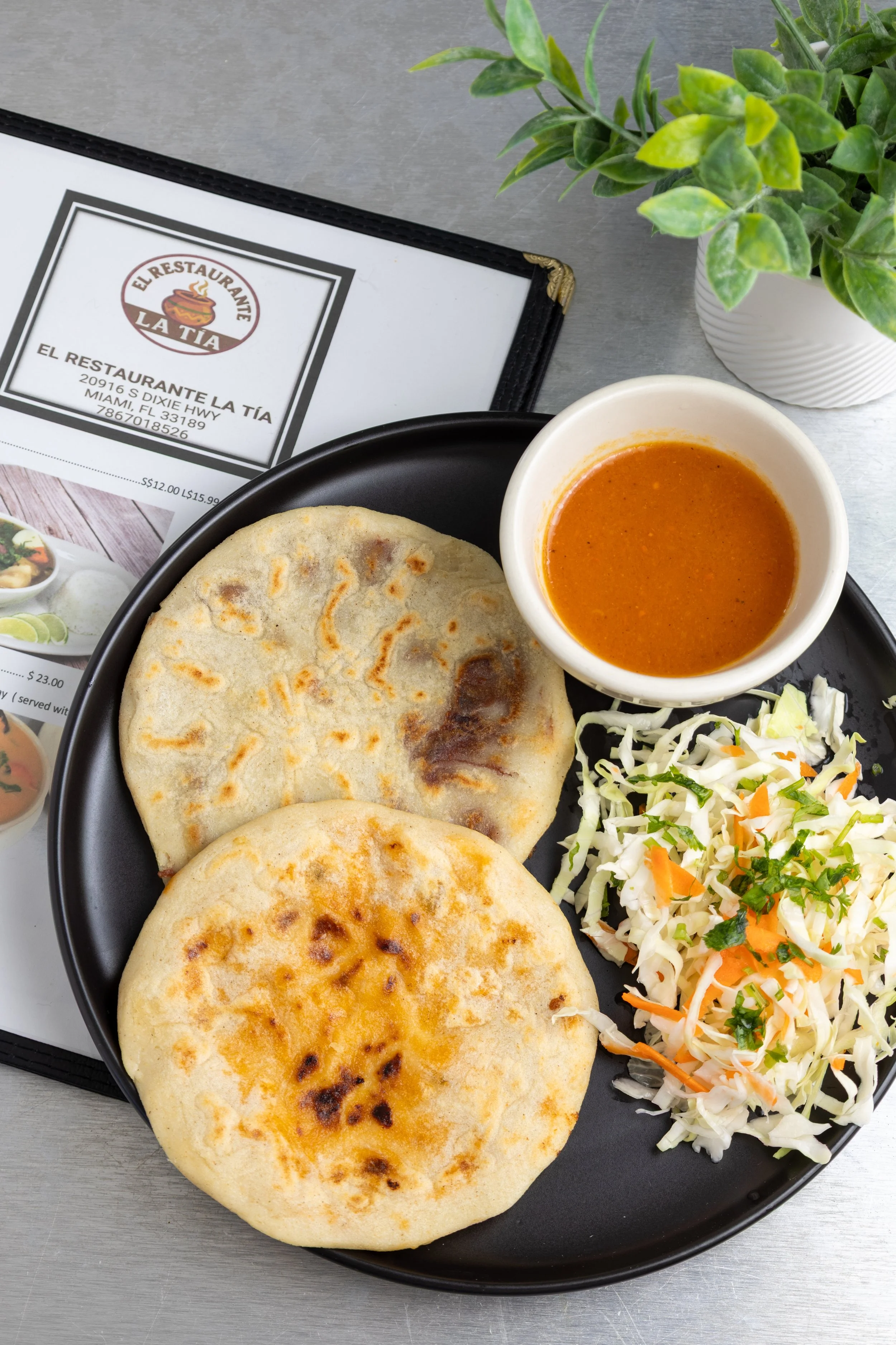 Plate with two stuffed flatbreads, a bowl of orange-colored sauce, and a small serving of shredded cabbage salad garnished with herbs, placed on a black round plate. Background includes a menu and a potted plant.