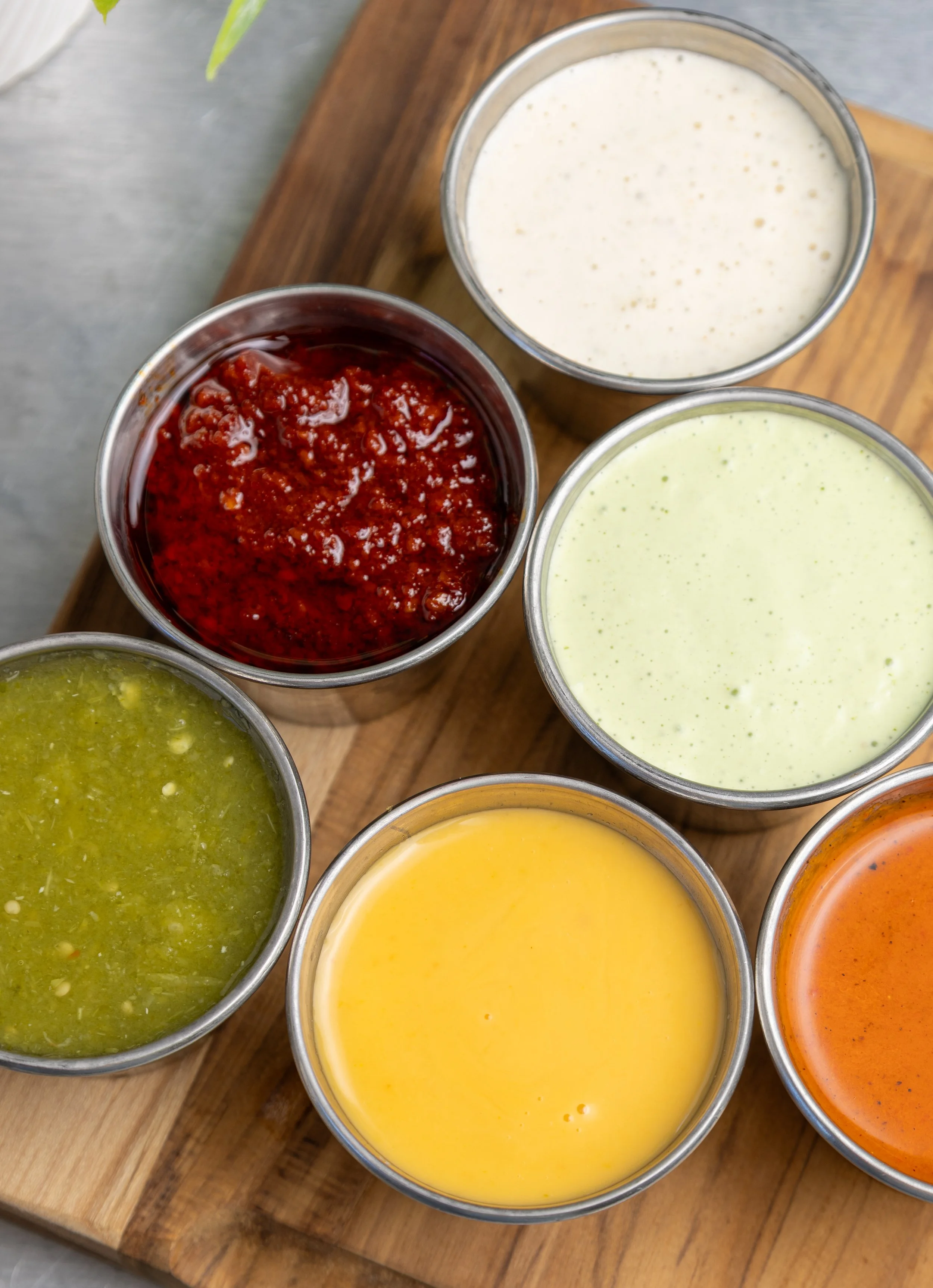 Six small glass bowls filled with various sauces or dressings, arranged on a wooden board. The sauces are red, green, yellow, orange, white, and off-white.
