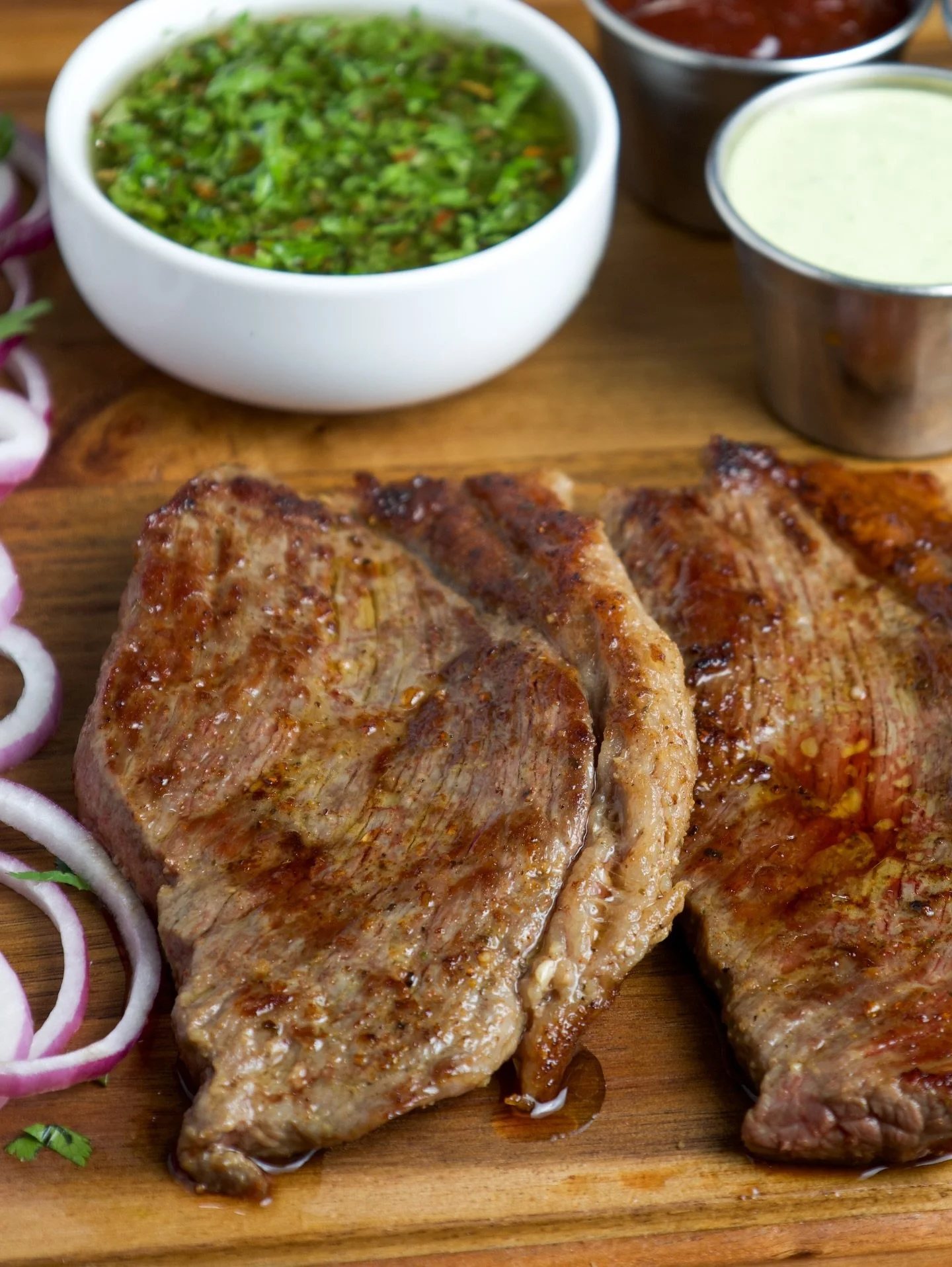 Two grilled steaks on a wooden cutting board with sliced red onions and cilantro, accompanied by bowls of green sauce, chopped herbs, and a creamy sauce.