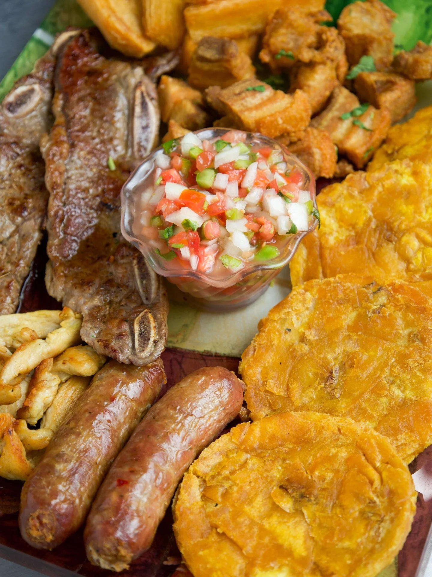 A platter of American foods including fried fish, sausage links, fried plantains, crispy fried pork belly pieces, a small bowl of pico de gallo, fried yuca, and fried sweet plantains.