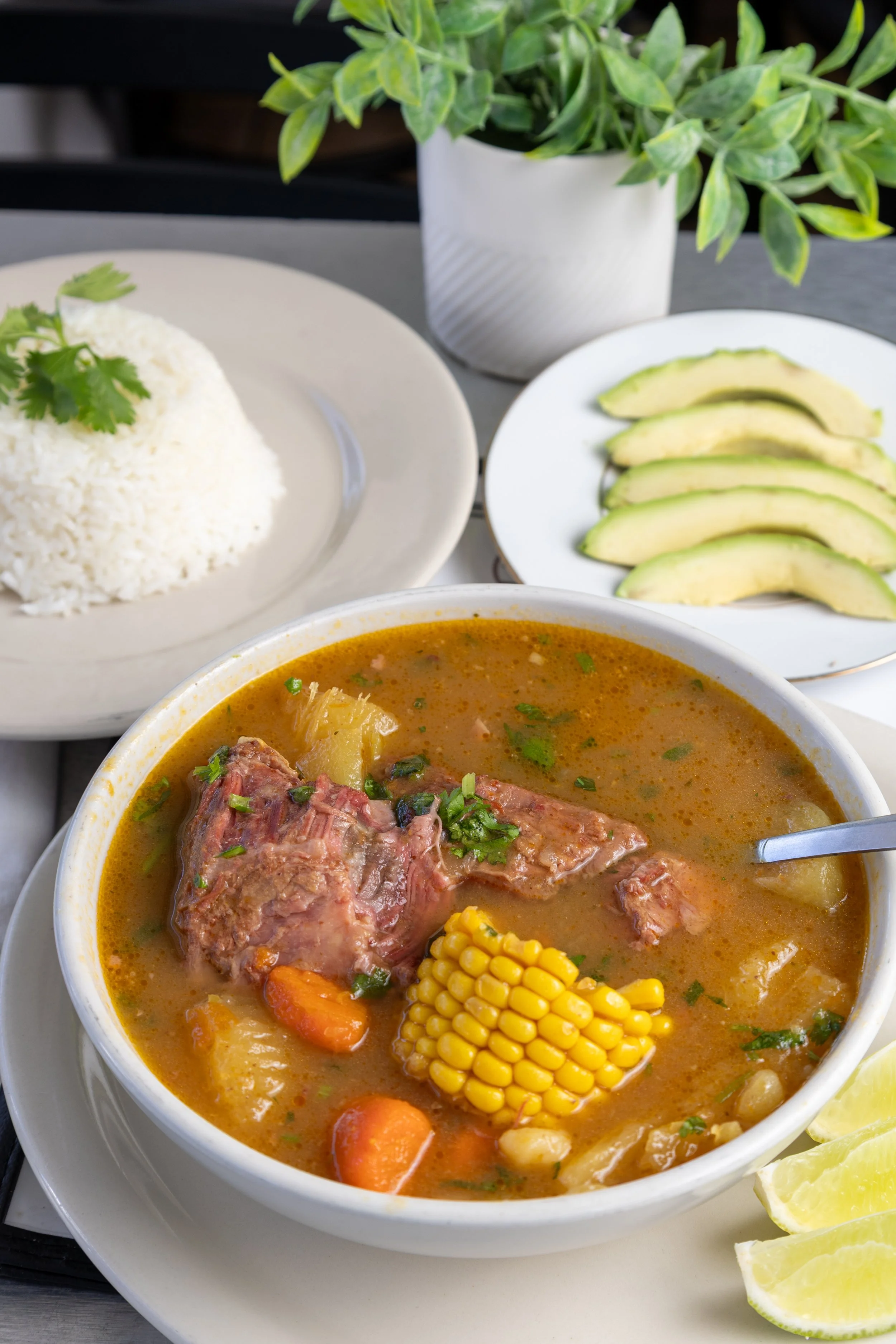 A bowl of beef stew with carrots, corn, and potatoes, served with lime wedges, a plate of sliced avocados, a plate of rice garnished with cilantro, and a potted green plant in the background.