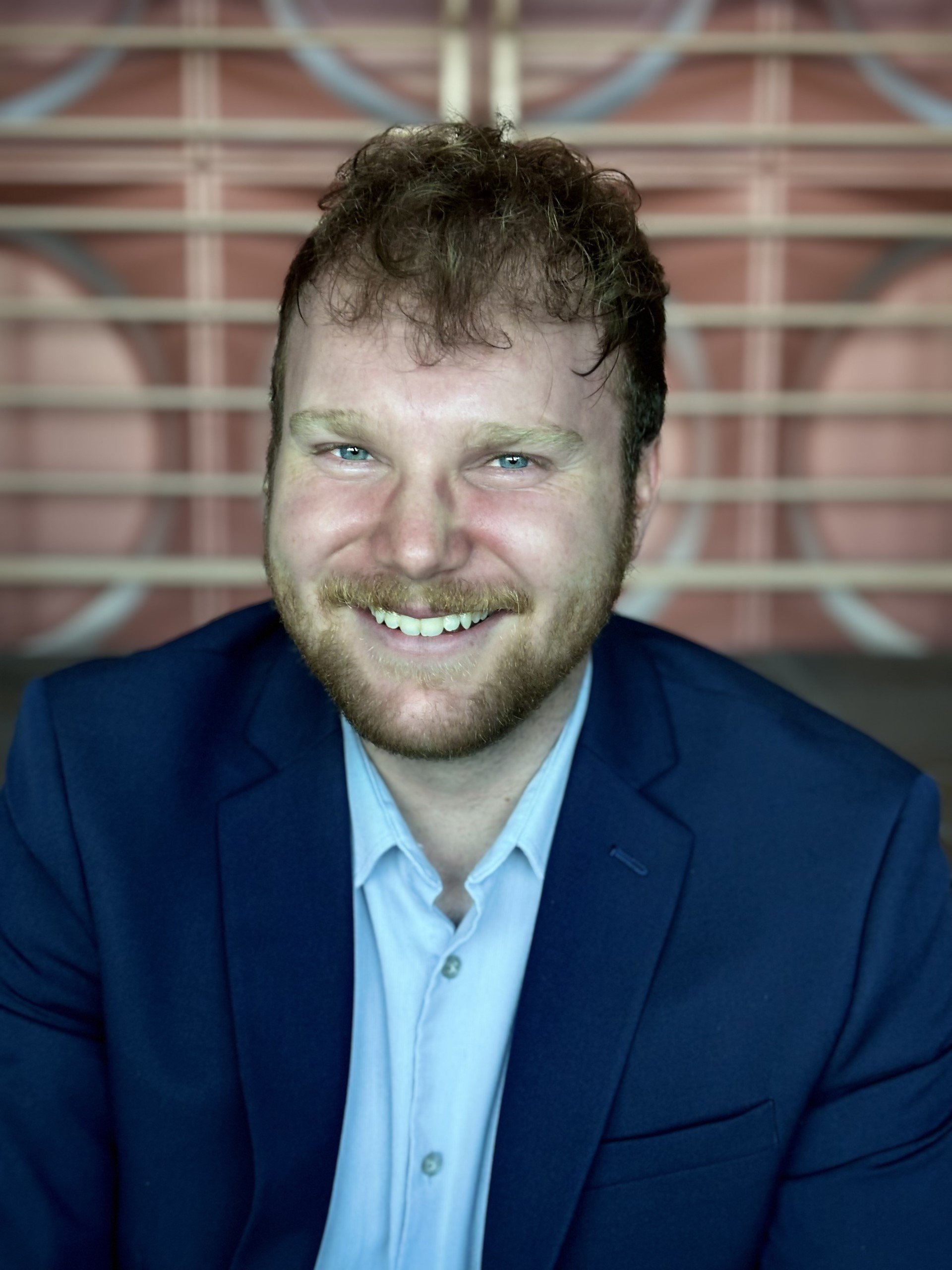 A man with short, curly brown hair, blue eyes, and a beard, smiling while wearing a blue suit jacket and a light blue dress shirt. The background features a geometric pattern with soft pink and beige tones.