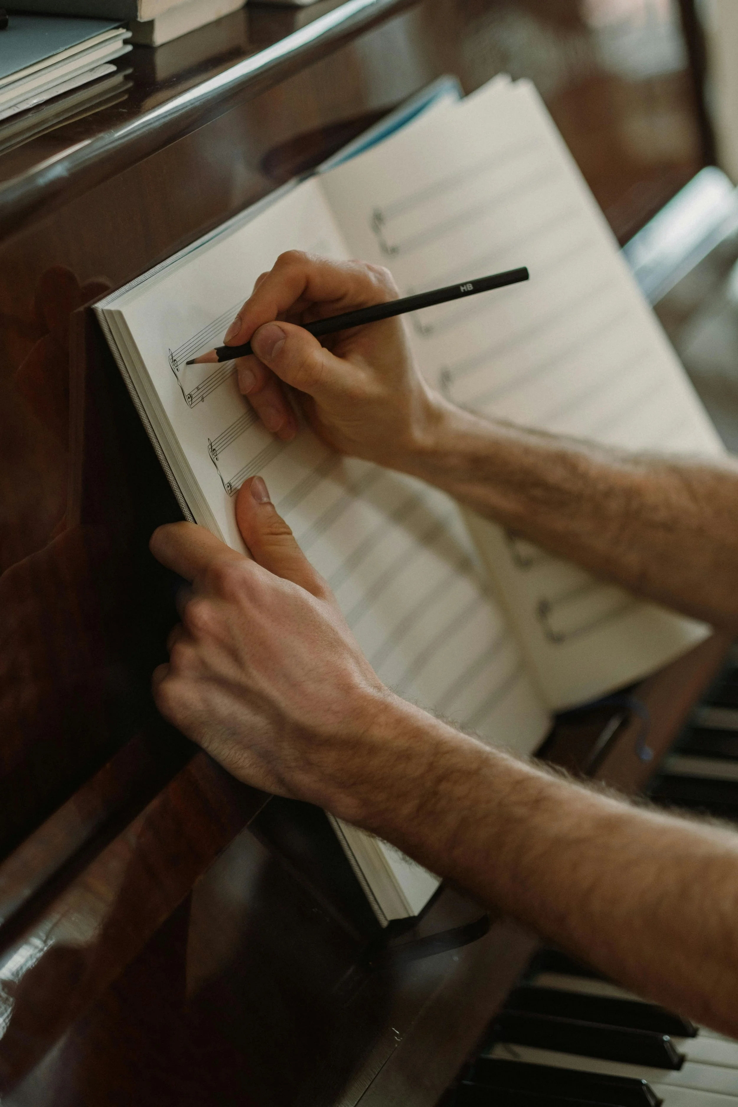 A person writing music notes in a music notebook with a pencil while sitting at a keyboard piano.
