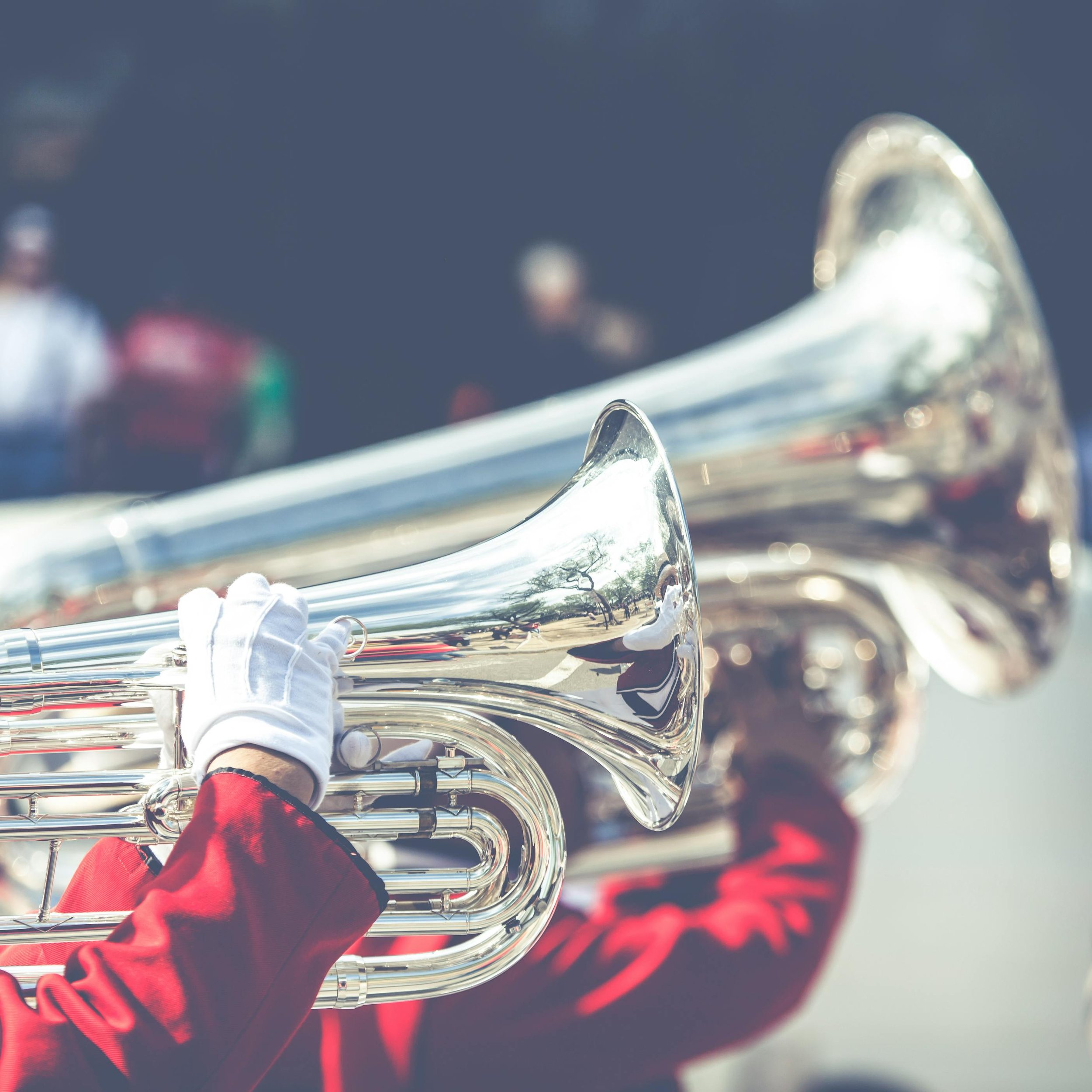 A person in a red jacket and white gloves playing a shiny silver sousaphone outdoors, with blurred figures in the background.