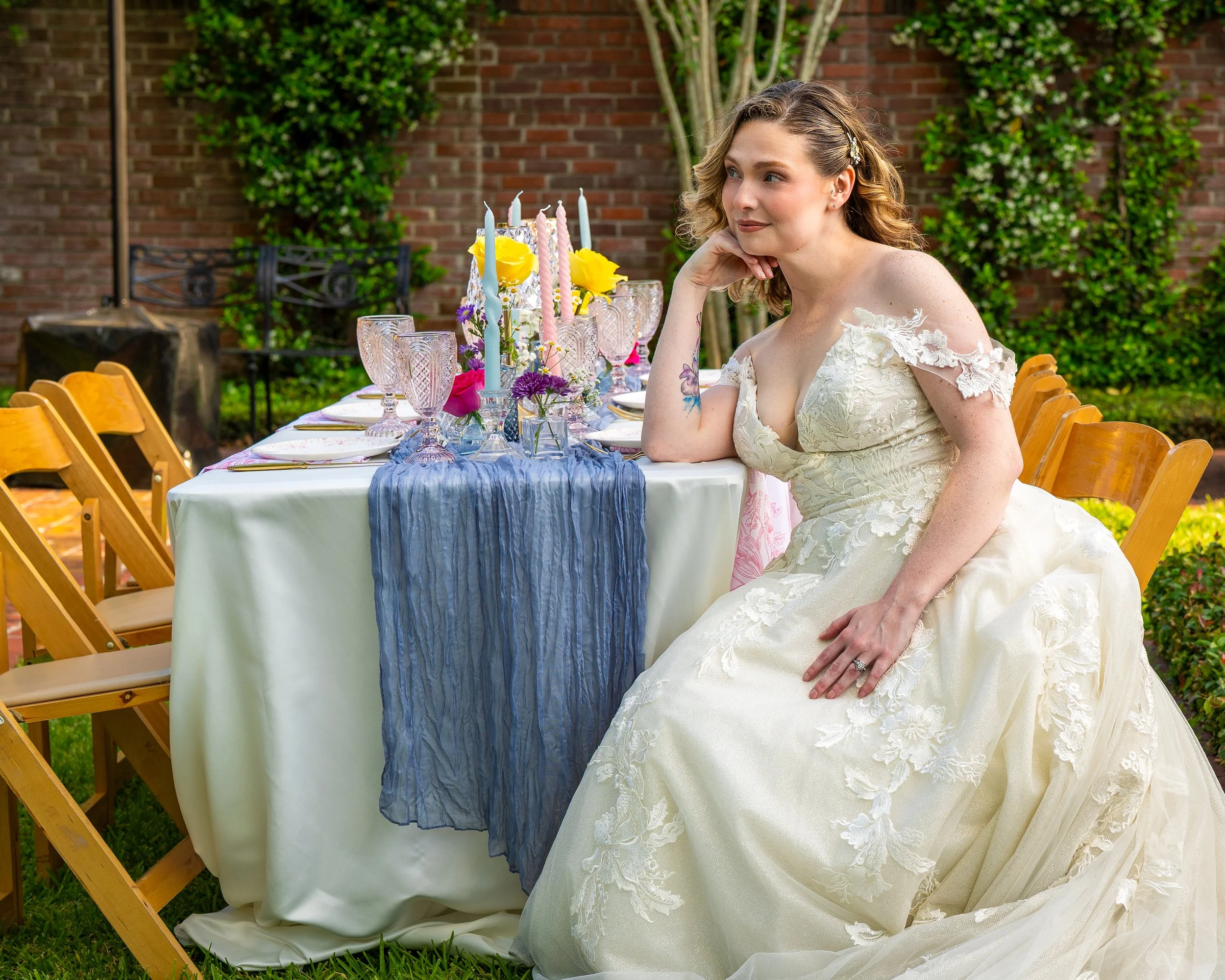 A woman in a wedding dress sitting at a decorated outdoor table, with a brick wall and green foliage in the background.