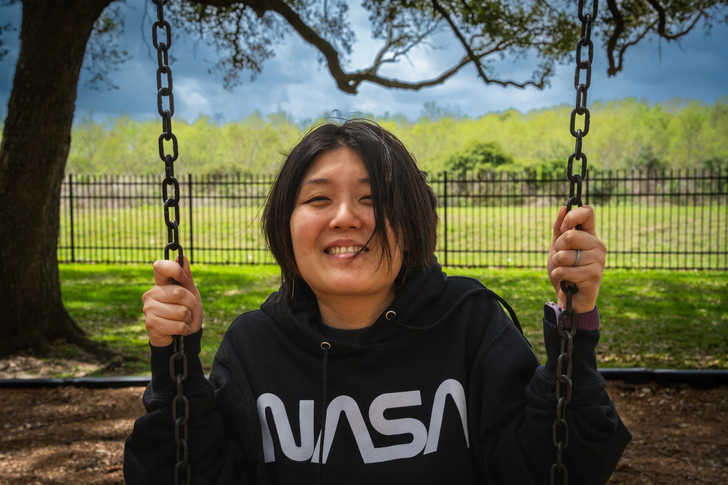 A woman with short dark hair smiling while sitting on a swing outdoors, with trees, a fence, and a cloudy sky in the background.