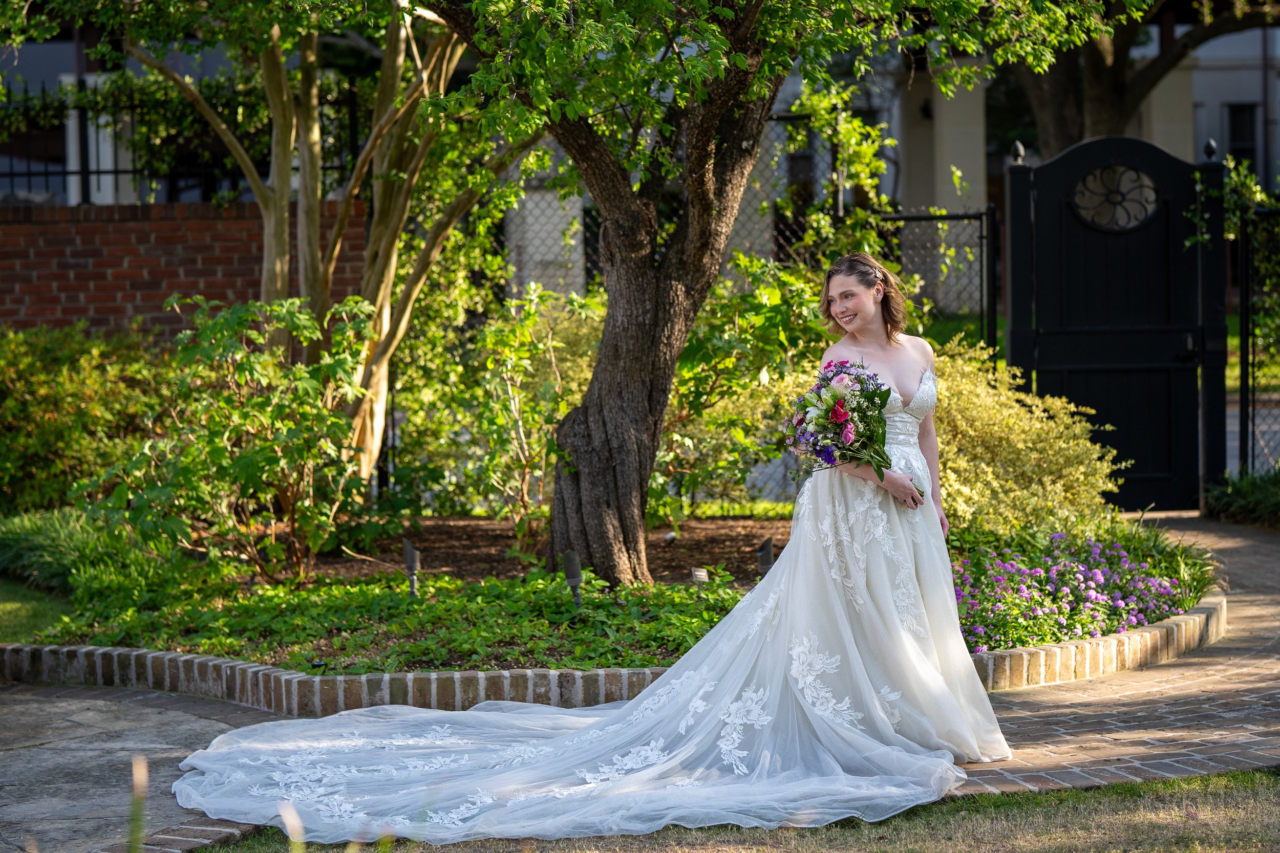 A woman in a white wedding gown holding a bouquet of flowers, standing outdoors in a garden area with trees, bushes, and a brick sidewalk.