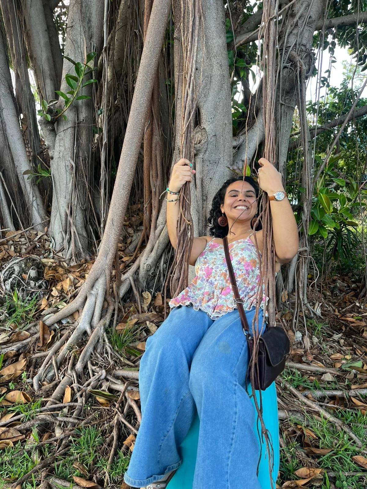 A woman sitting on a swing made of tree roots and vines in front of a large tree with roots and vines. She is smiling and looking at the camera, wearing a floral top, blue jeans, and accessories. The background features green foliage and the ground covered in dry leaves.
