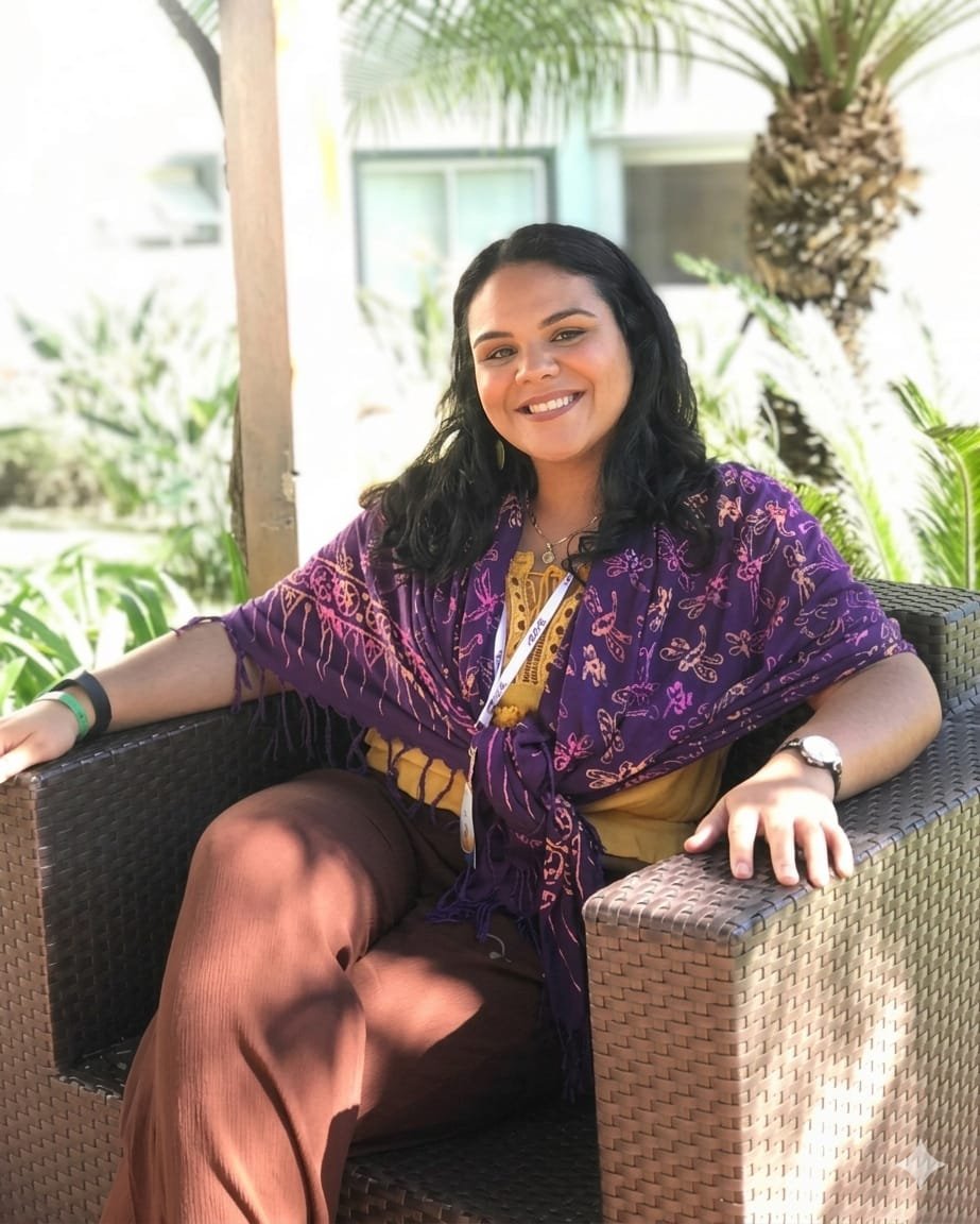 A woman with black, curly hair sitting on a wicker sofa, smiling at the camera, with greenery and palm trees in the background.