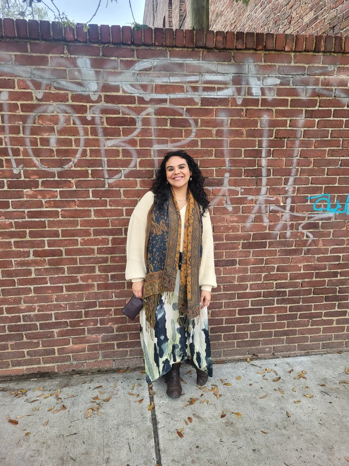 A woman with dark curly hair and a bright smile standing in front of a brick wall with graffiti. She is wearing a cream-colored sweater, a multi-colored patterned skirt, and a long scarf with intricate designs. She is holding a small dark clutch purse and wearing brown boots. There are fallen leaves on the sidewalk.