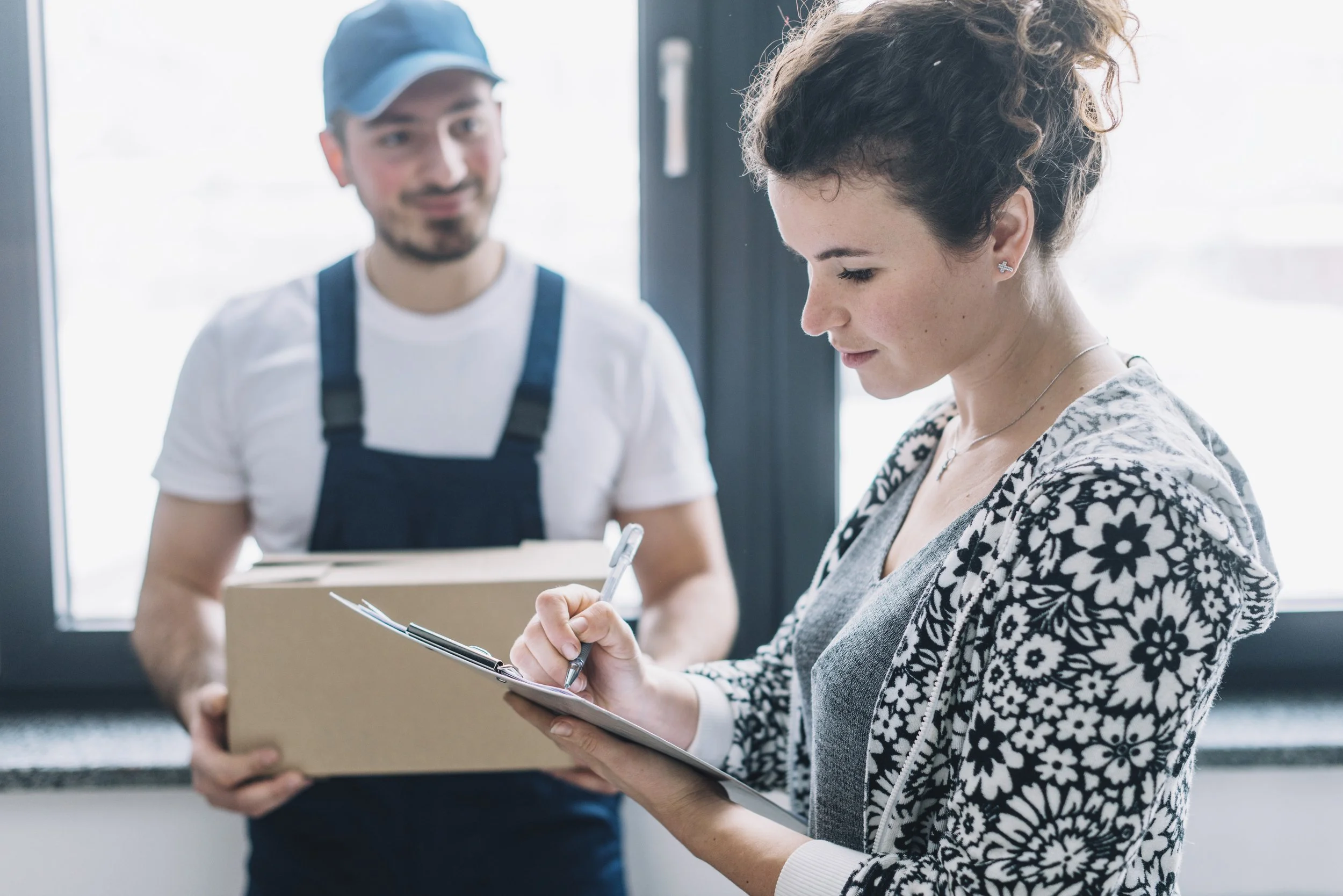 A woman with short curly hair wearing a floral jacket writes in a notebook while a man in a baseball cap and apron holds a box in the background.