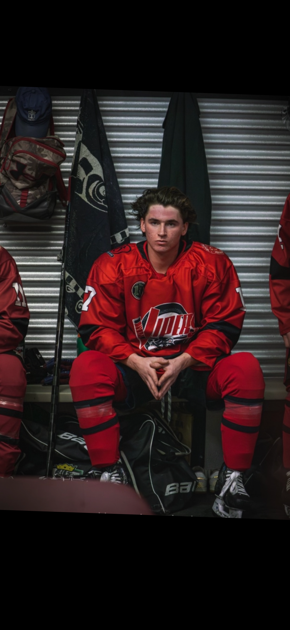 Young male hockey player with long hair, sitting in a locker room, wearing a red hockey jersey with a logo that says 'Vipers.' He is flanked by other teammates, with gear around his feet and hanging on hooks behind him.