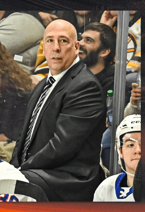 A man in a black suit and tie standing courtside at a hockey game, with a serious expression, surrounded by spectators and a hockey player.