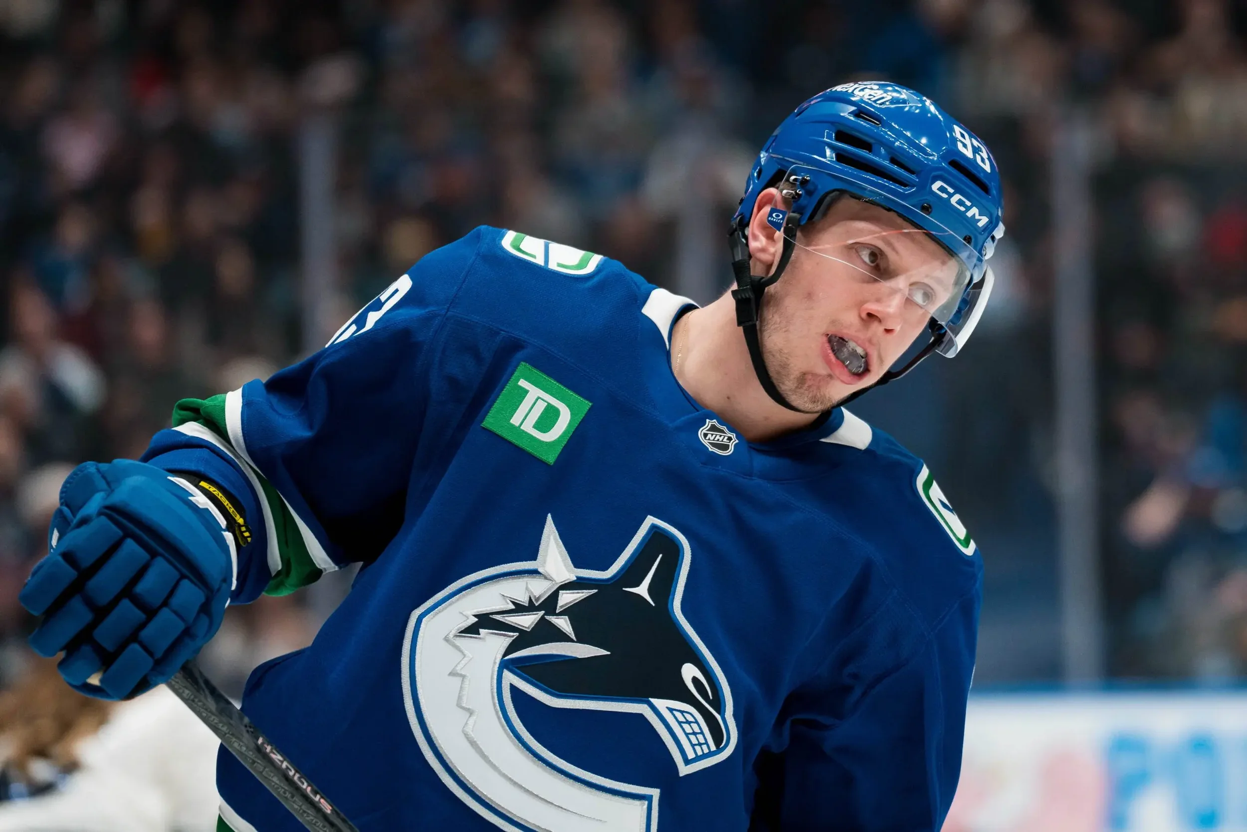 A male hockey player wearing a blue jersey and helmet, skating during a game, with a crowd in the background.