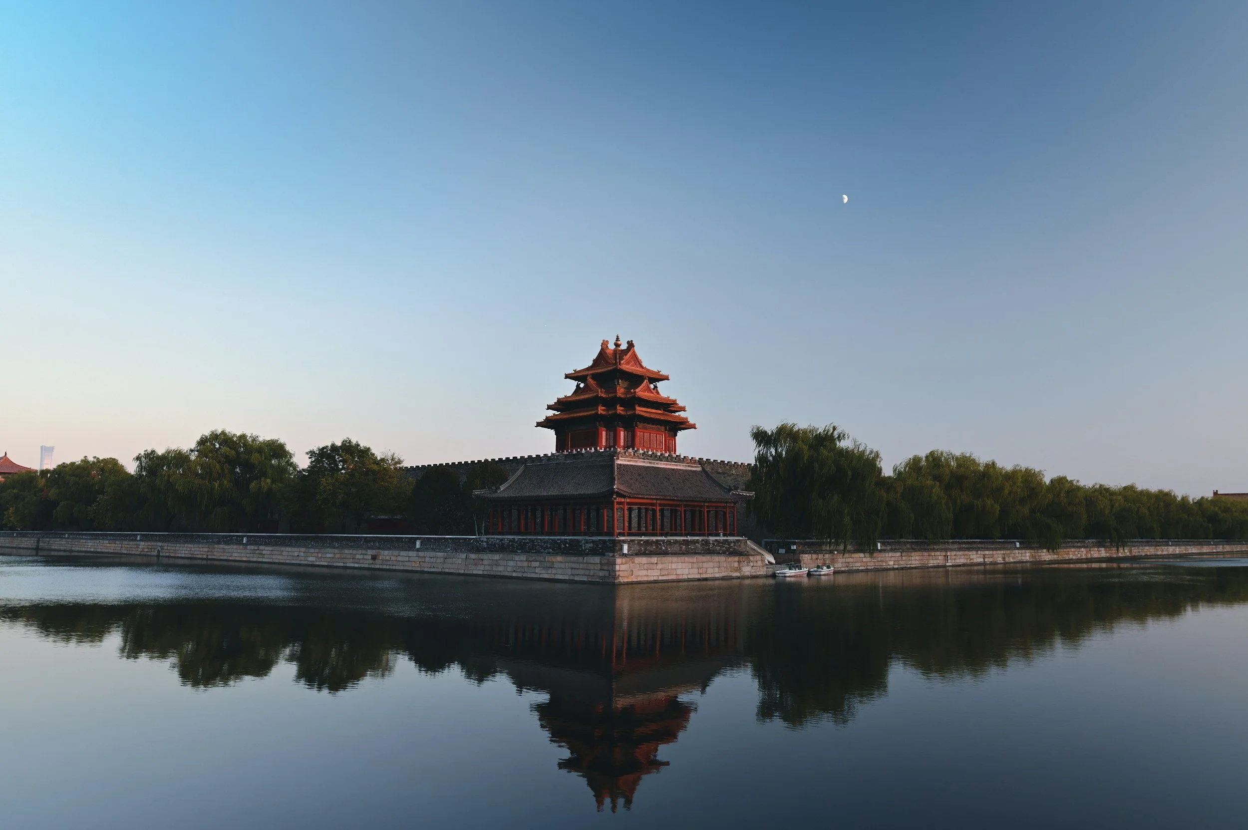 Traditional Chinese-style building beside a body of water, with trees and a clear sky in the background, and a crescent moon visible in the sky.