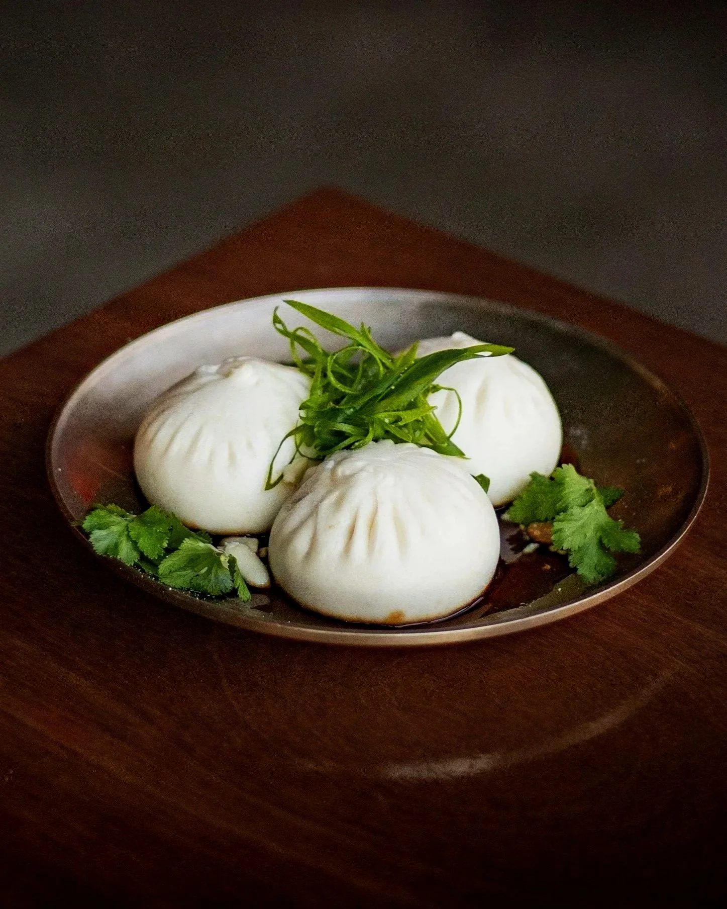 Four steamed Chinese dumplings on a metal plate garnished with green herbs and cilantro leaves, placed on a wooden surface.